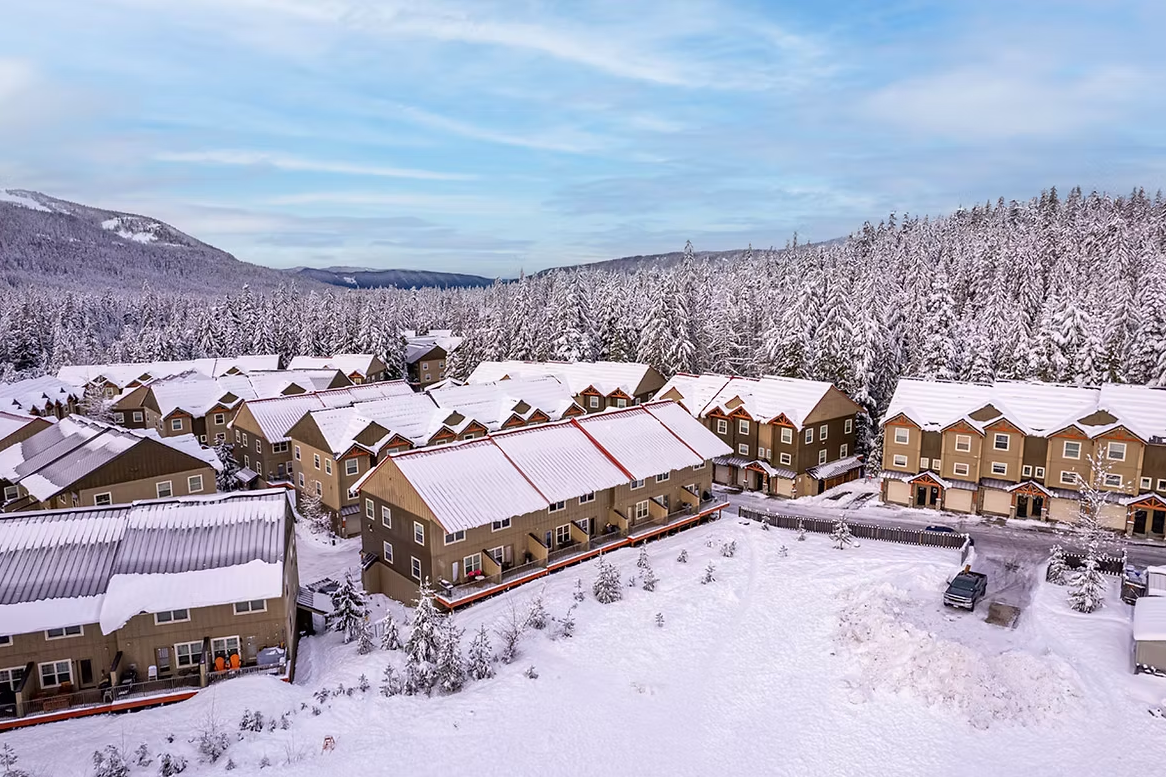 A snowy mountain landscape with a cluster of beige apartment buildings with snow-covered roofs, in front of a forest of snow-covered pine trees and mountains in the background.