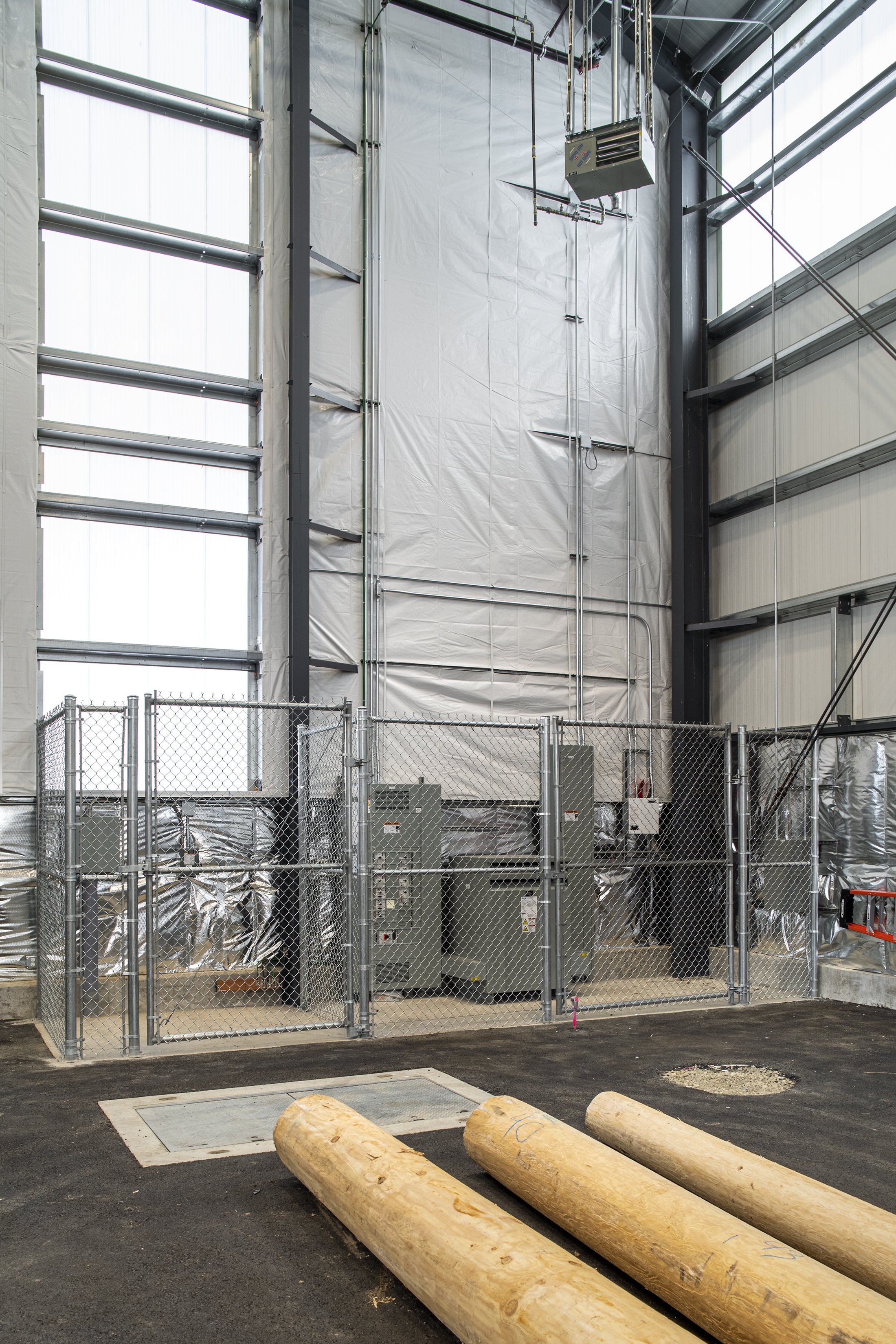 Inside a construction or industrial building exposing large electrical panels, wiring, and ductwork, with two wooden logs on the ground.