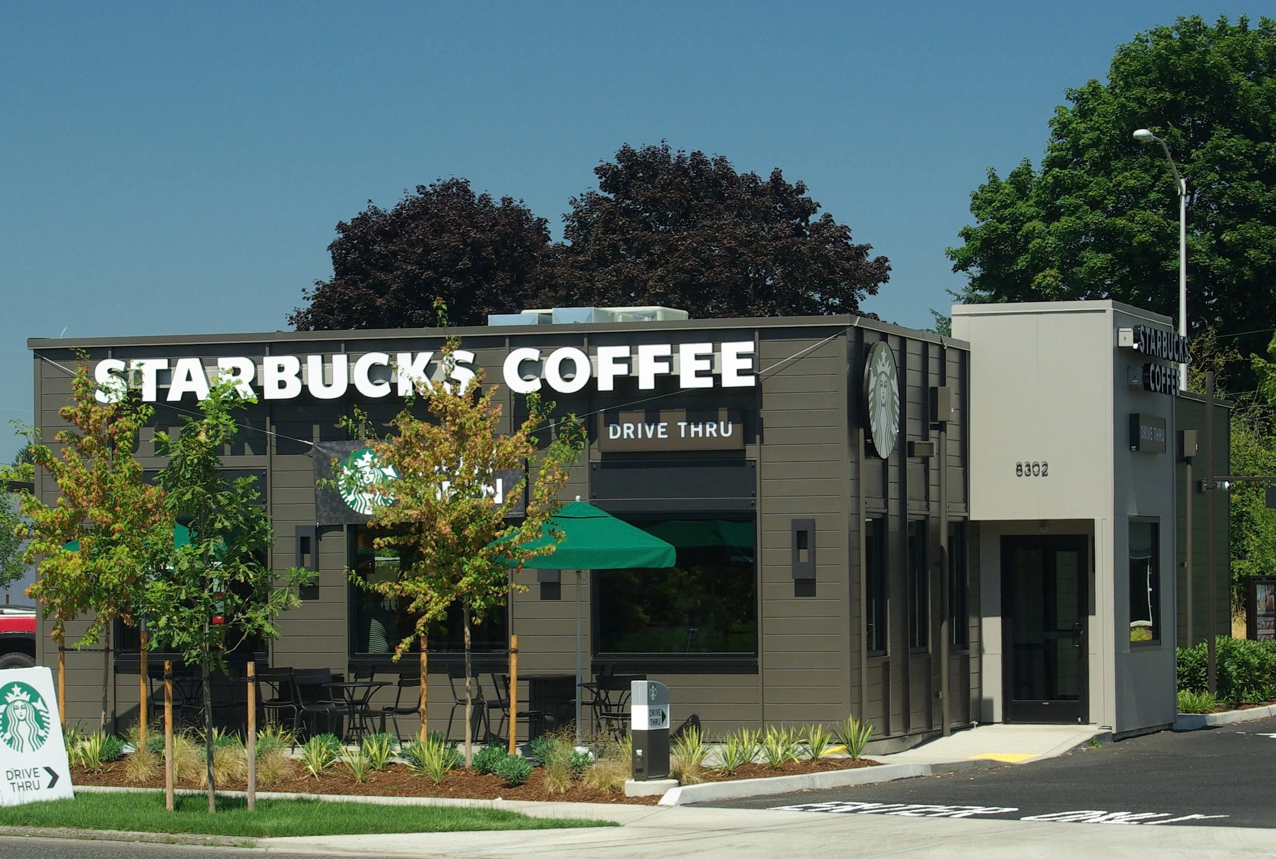 Starbucks Coffee shop with a drive-thru sign, outdoor seating, trees, and a parking lot.