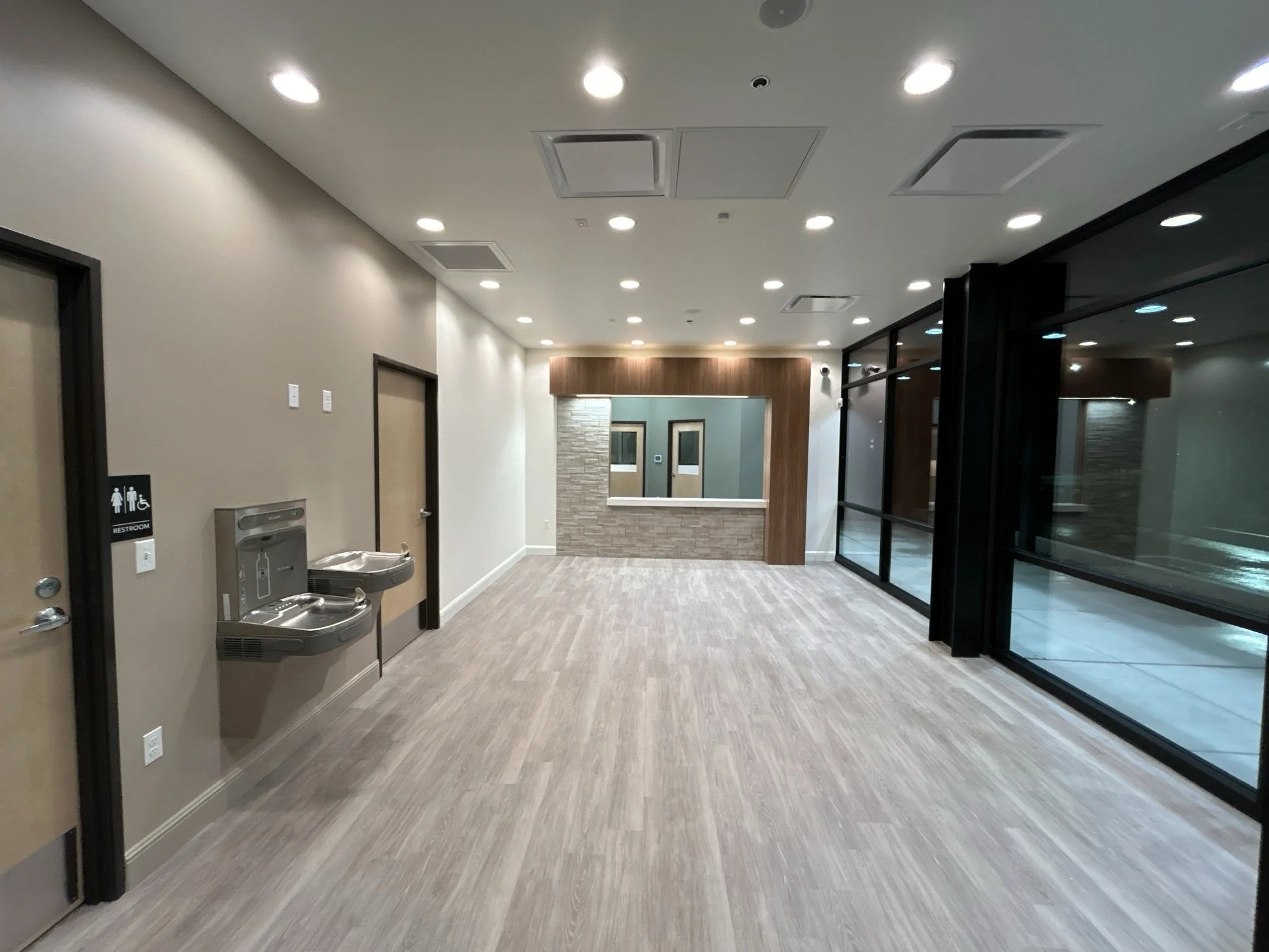 Empty interior space with beige walls, wooden floor, and large glass windows on the right. Ceiling has recessed lighting, and there is a water fountain and restroom sign on the left wall. A reception window with a wooden frame is visible in the backg