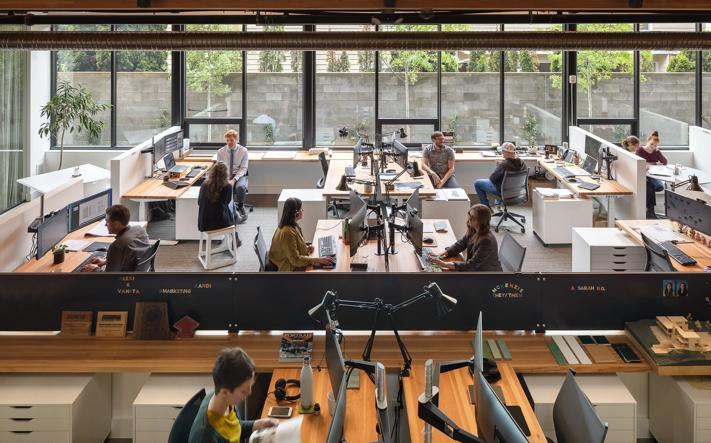 An open-plan modern office with multiple employees working at desks equipped with monitors and computers. Large windows provide natural light, and there are potted plants and personal items on desks. The office has a contemporary design with wooden d