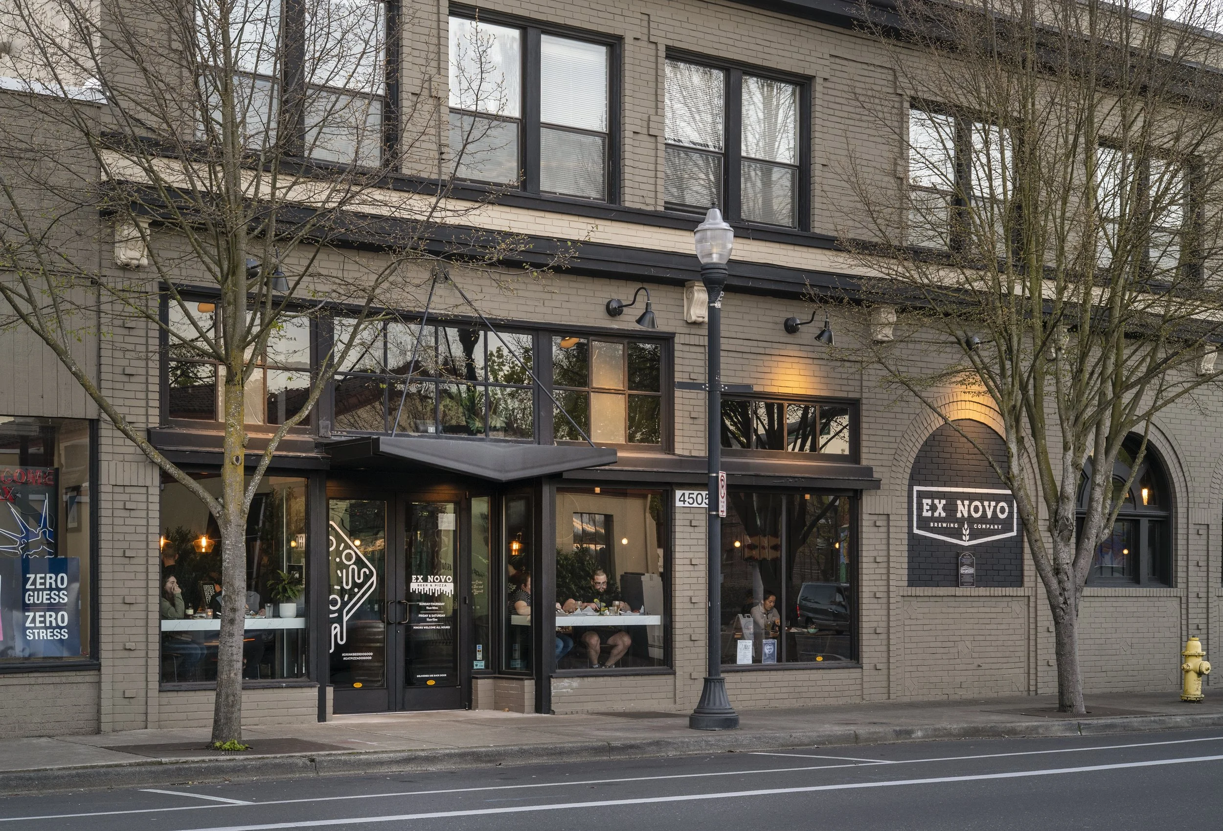 Exterior of Ex Novo Brewing Company with large windows, brick facade, trees, and streetlamp in front.