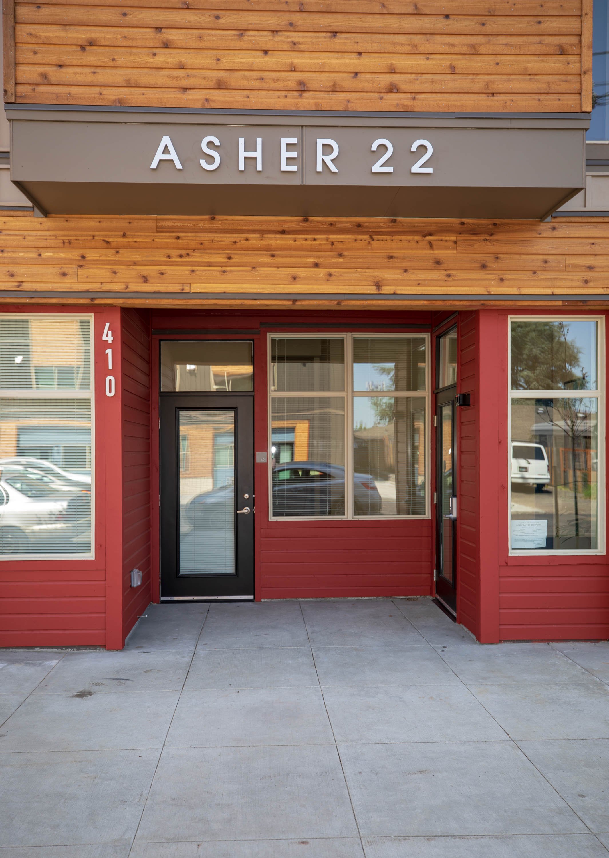 Exterior of a commercial building with a wooden upper facade, a gray sign reading 'ASHER 22,' and a red lower facade with large windows and a black door.