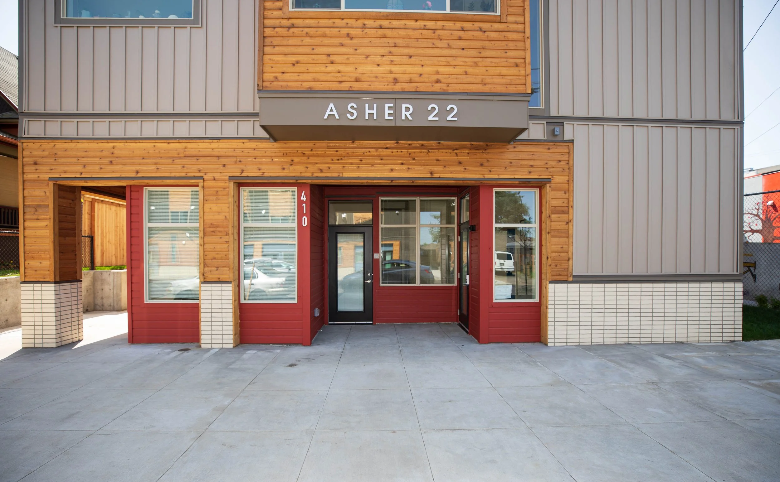 Modern residential building with wooden and gray exterior, red accents around the entrance, and a sign that reads 'ASHER 22' above the doorway.