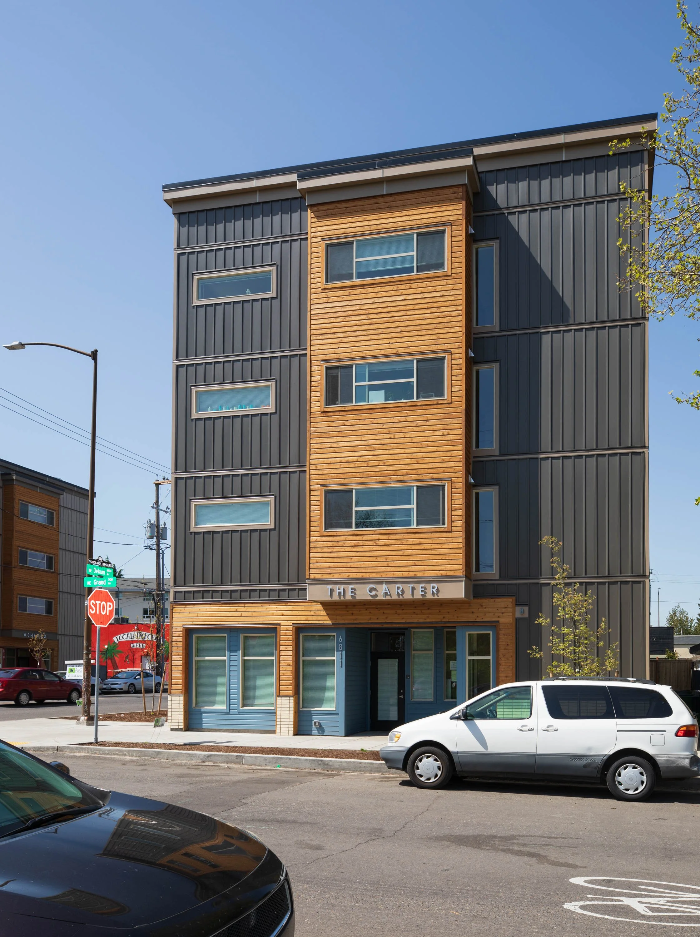 A modern multi-story apartment building with a wooden and dark metal exterior, labeled "The Carter," located on a city street corner with cars parked in front and a stop sign visible.