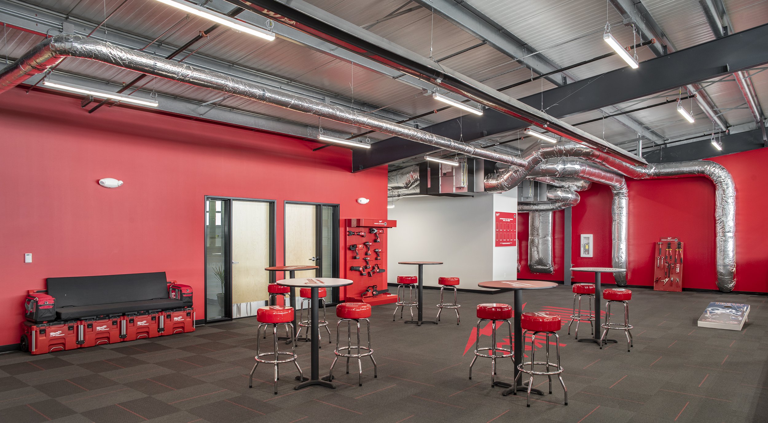 An industrial style break room with red walls, silver ventilation ducts, and black ceiling. There are high tables with red cushioned stools, a black seating bench with toolboxes, and a pegboard with tools on the wall. The floor is carpeted in dark gr