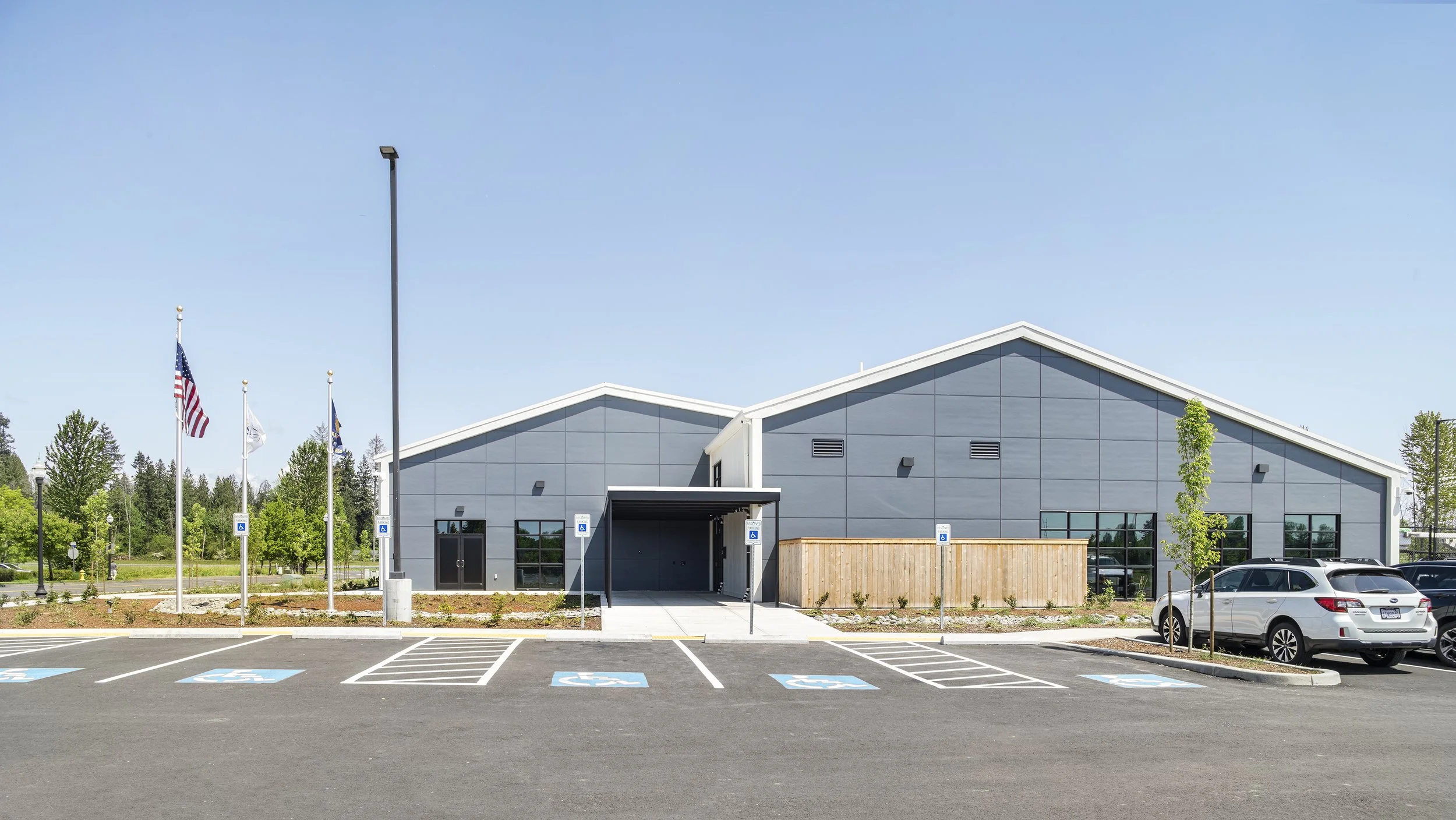 Modern grey building with a parking lot in front, designated for handicap parking. American flags and flagpoles are in the background, with trees and blue sky.