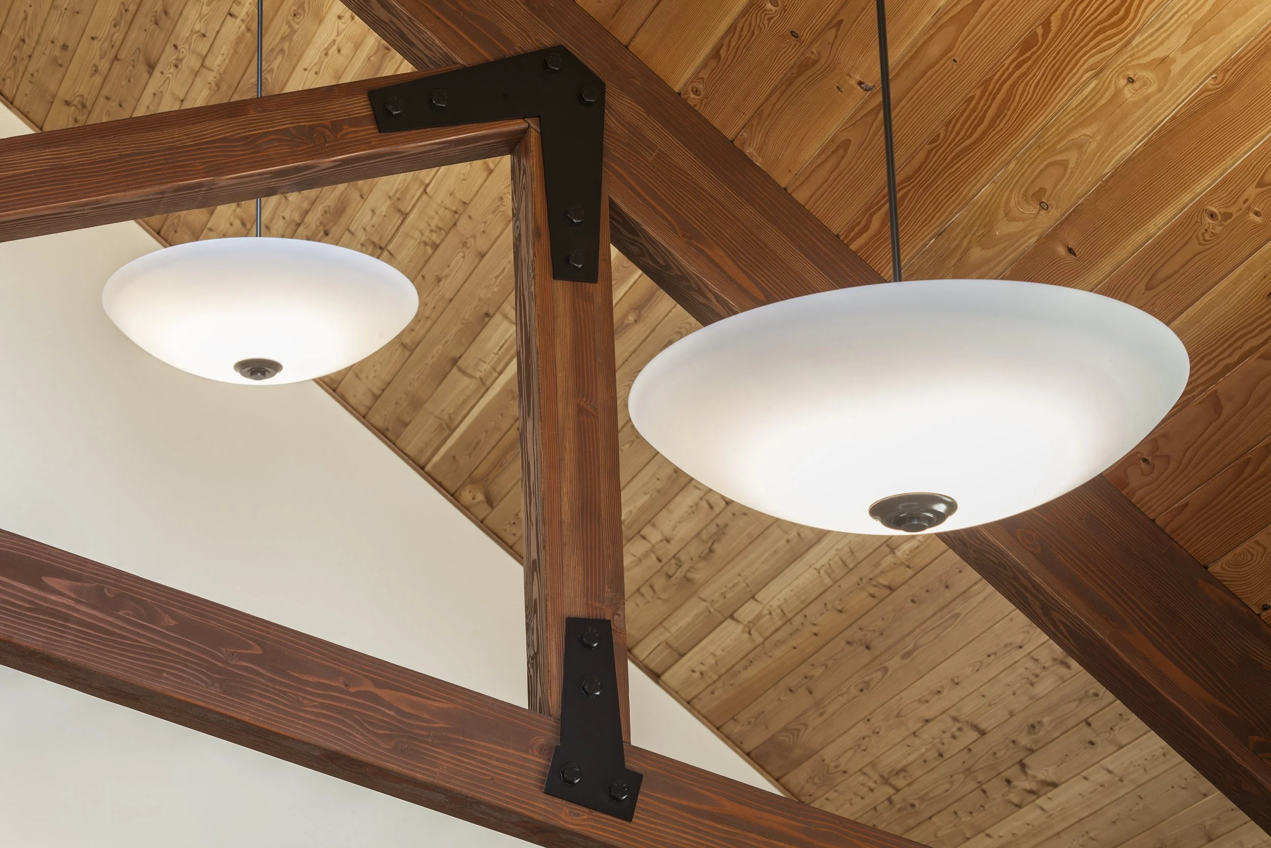 Two ceiling lights, one smaller and one larger, hanging from a ceiling with wood paneling, and a wooden structure with black metal brackets.