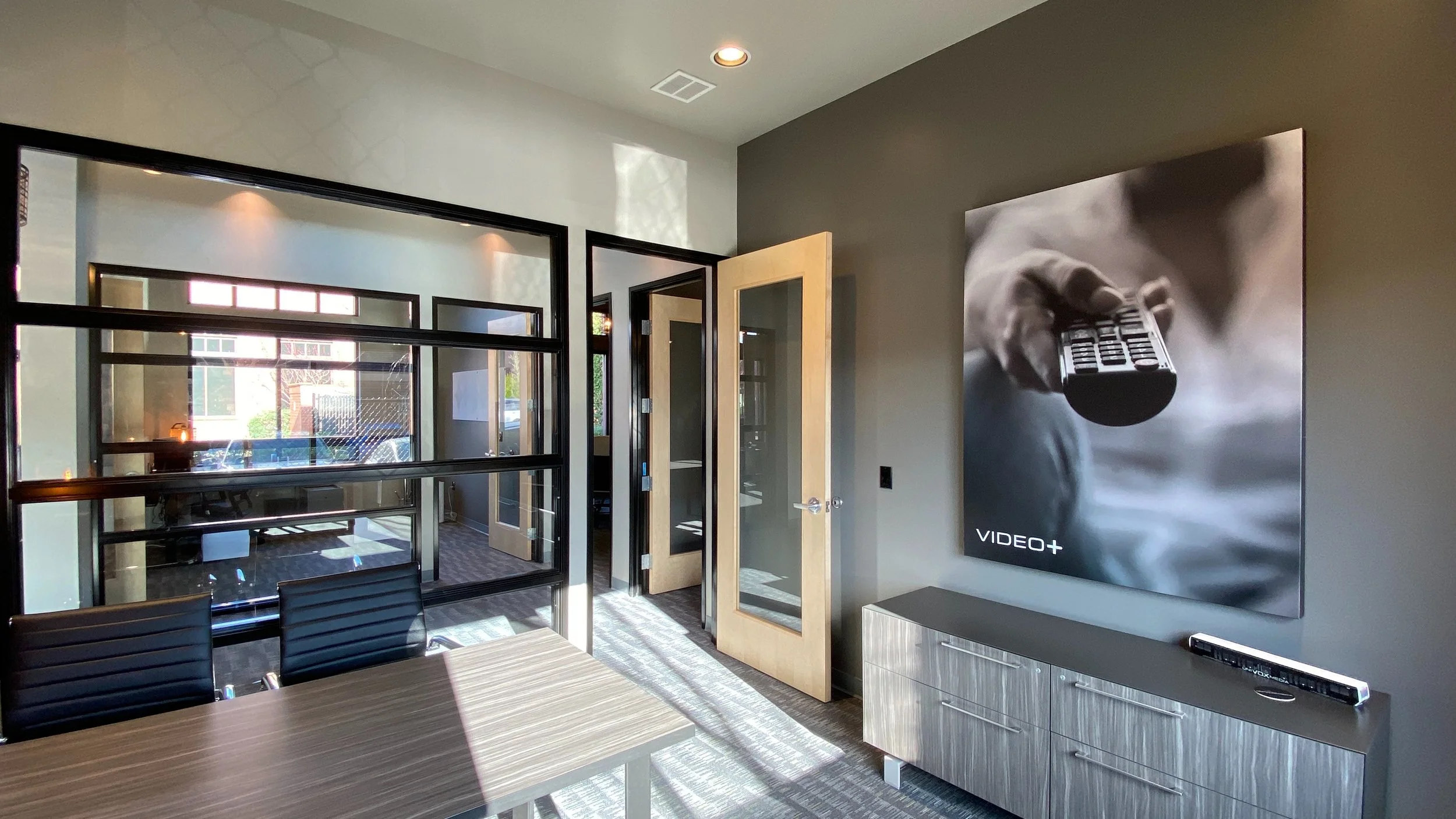 Modern office conference room with wooden table, black chairs, glass walls, large TV screen displaying "Video+" logo, and a soundbar on a light gray cabinet.