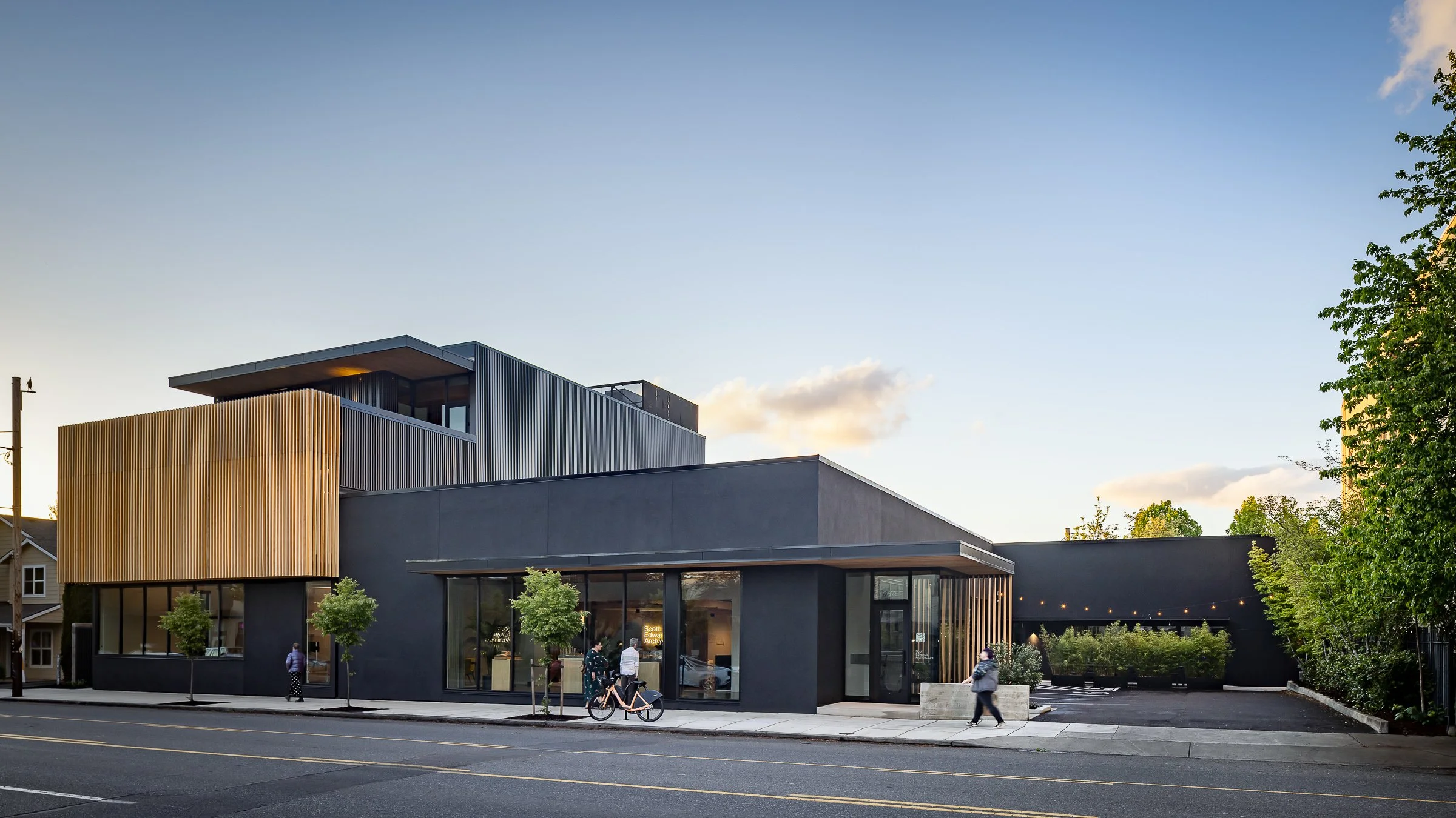 Modern multi-story building with black and wooden exterior, large glass windows, and small trees on the sidewalk, during daytime with clear sky.