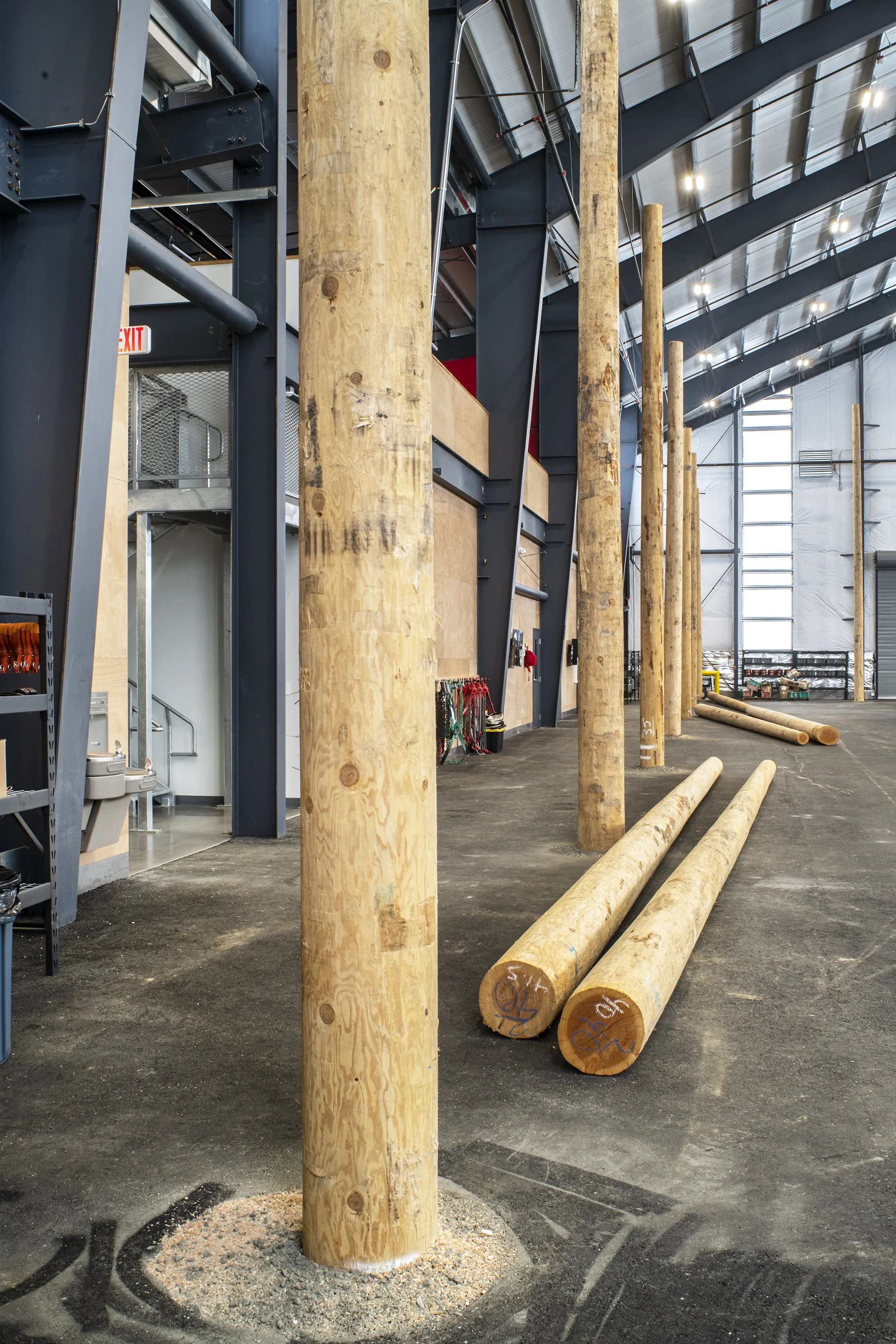 Several wooden poles are arranged vertically on a dark indoor gym floor, with some lying on the ground. The setting appears to be a warehouse or training facility with industrial metal beams and a high ceiling with bright lighting.