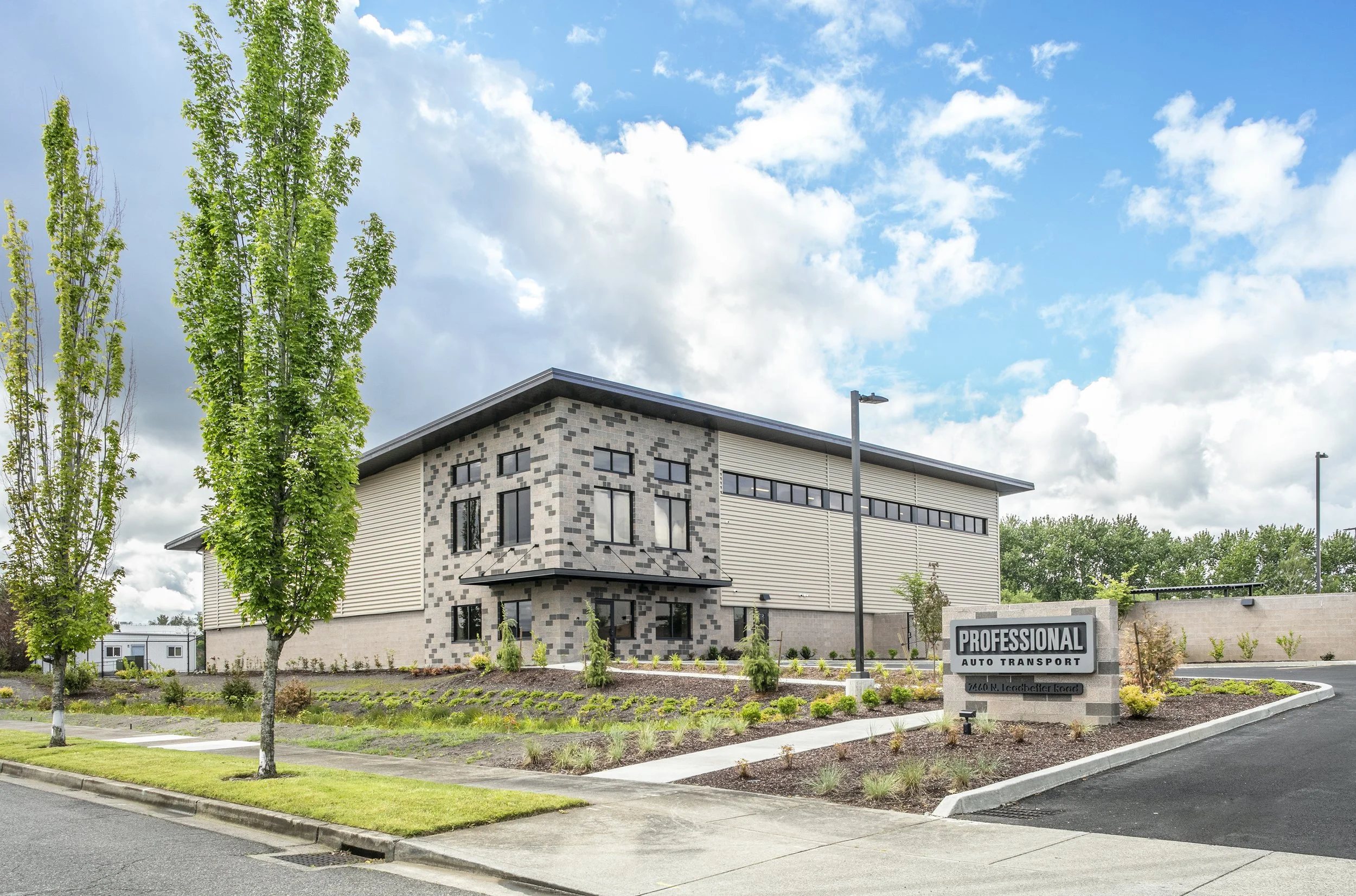 Modern commercial building with landscaped front yard and parking lot, sign reading 'Professional Auto Transport,' under partly cloudy sky.