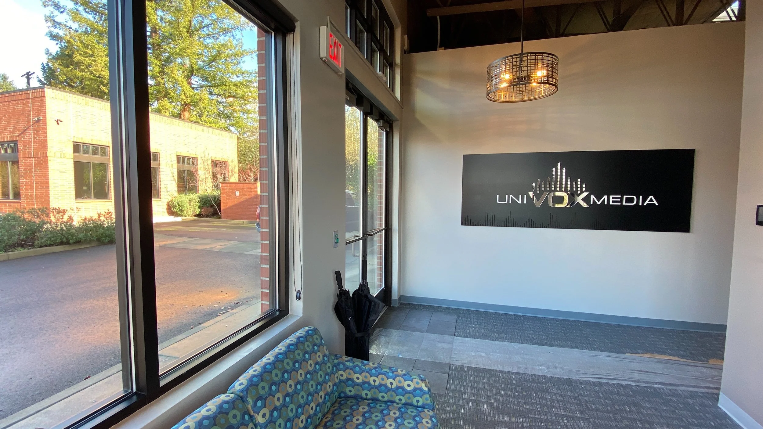 Interior of a reception area with large windows, a patterned couch, umbrella stand, and a wall sign reading 'Unity Vox Media' in a modern office building.