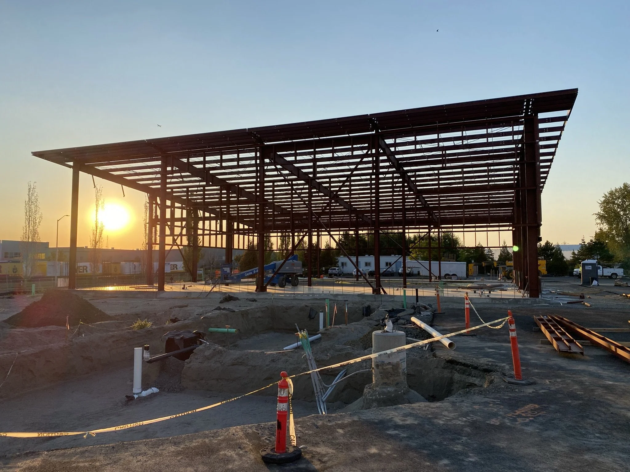Construction site with a steel framework for a new building, sunset in the background, construction equipment, pipes, and safety cones.