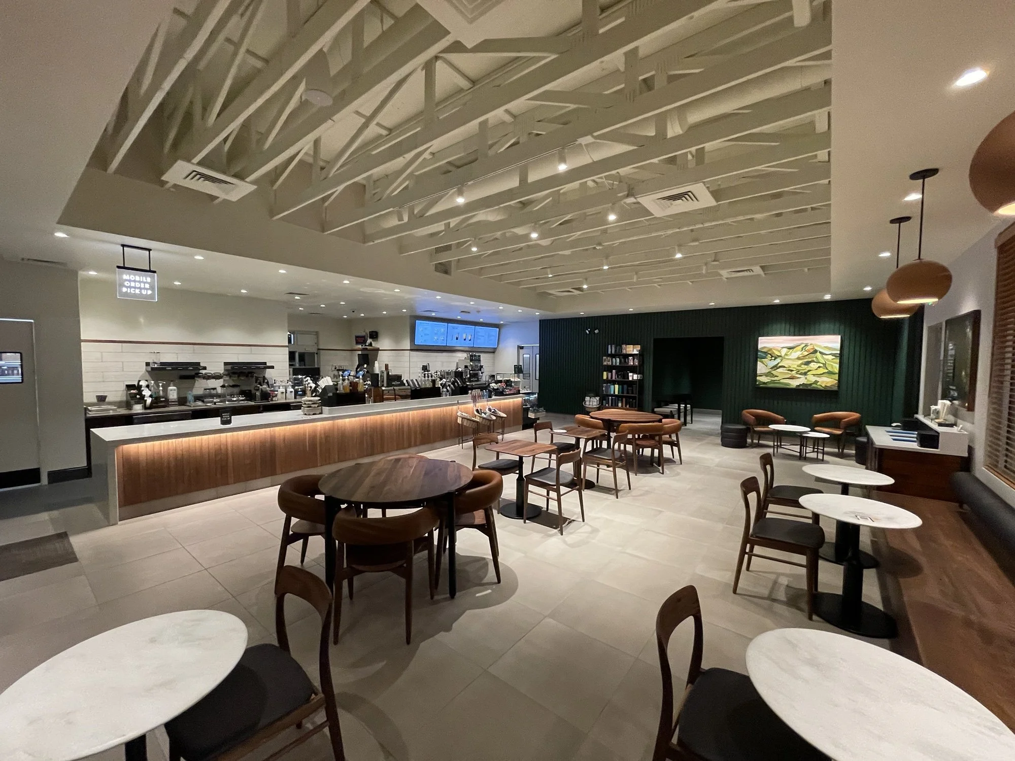 A modern, well-lit coffee shop with a counter on the left side, featuring coffee machines and a digital menu screen. The seating area includes white and dark wood tables with matching chairs, some with black cushions. There are pendant lights hanging