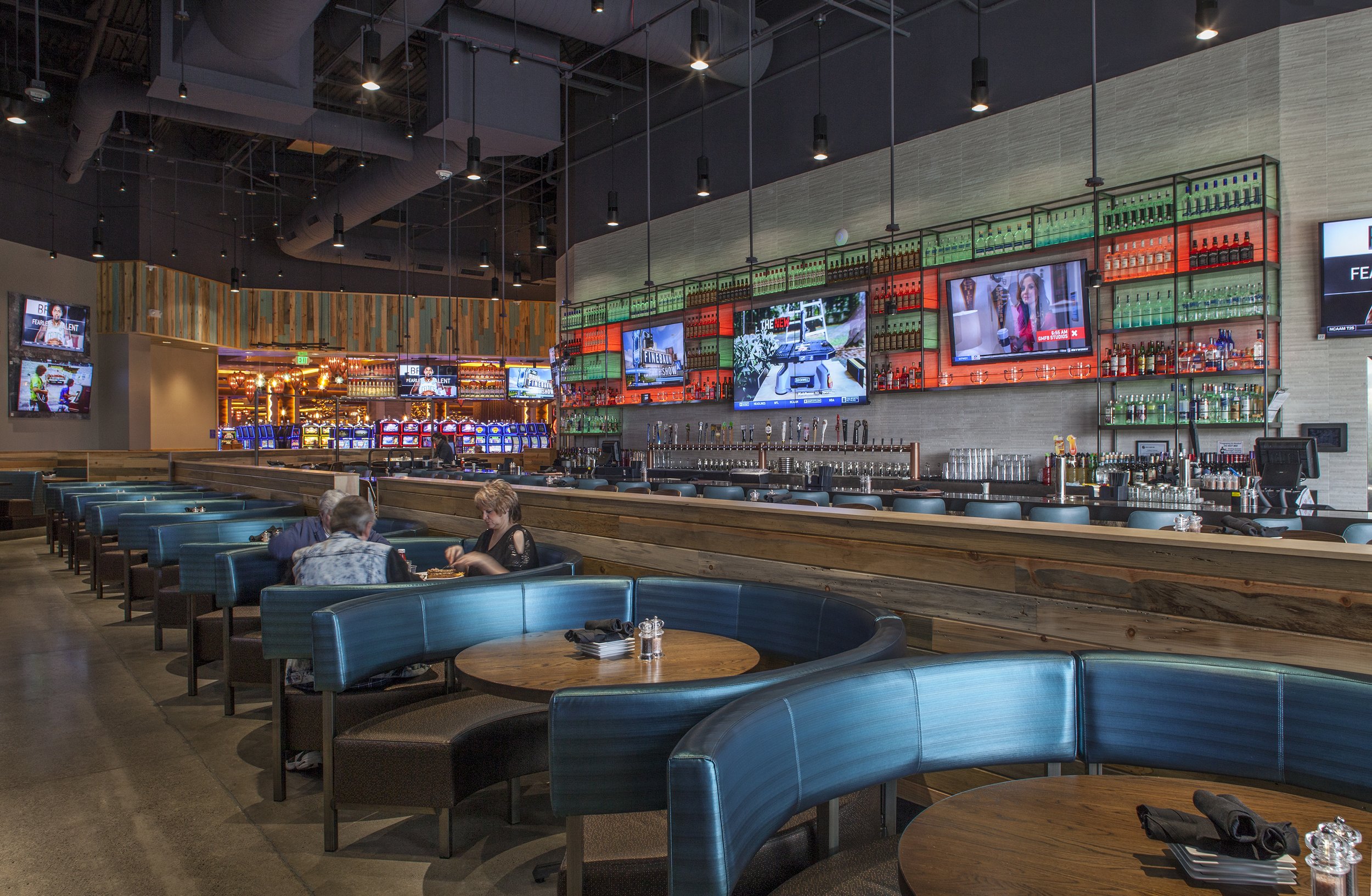 Interior of a modern bar or restaurant with blue booth seating, a long bar counter with multiple beer taps, several electronic screens on the wall displaying images and ads, and some people sitting at the booths.