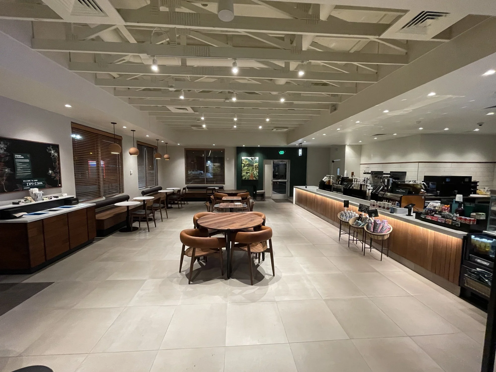 Empty modern coffee shop with wooden tables and chairs, a counter with coffee machines, and large windows with wooden blinds.