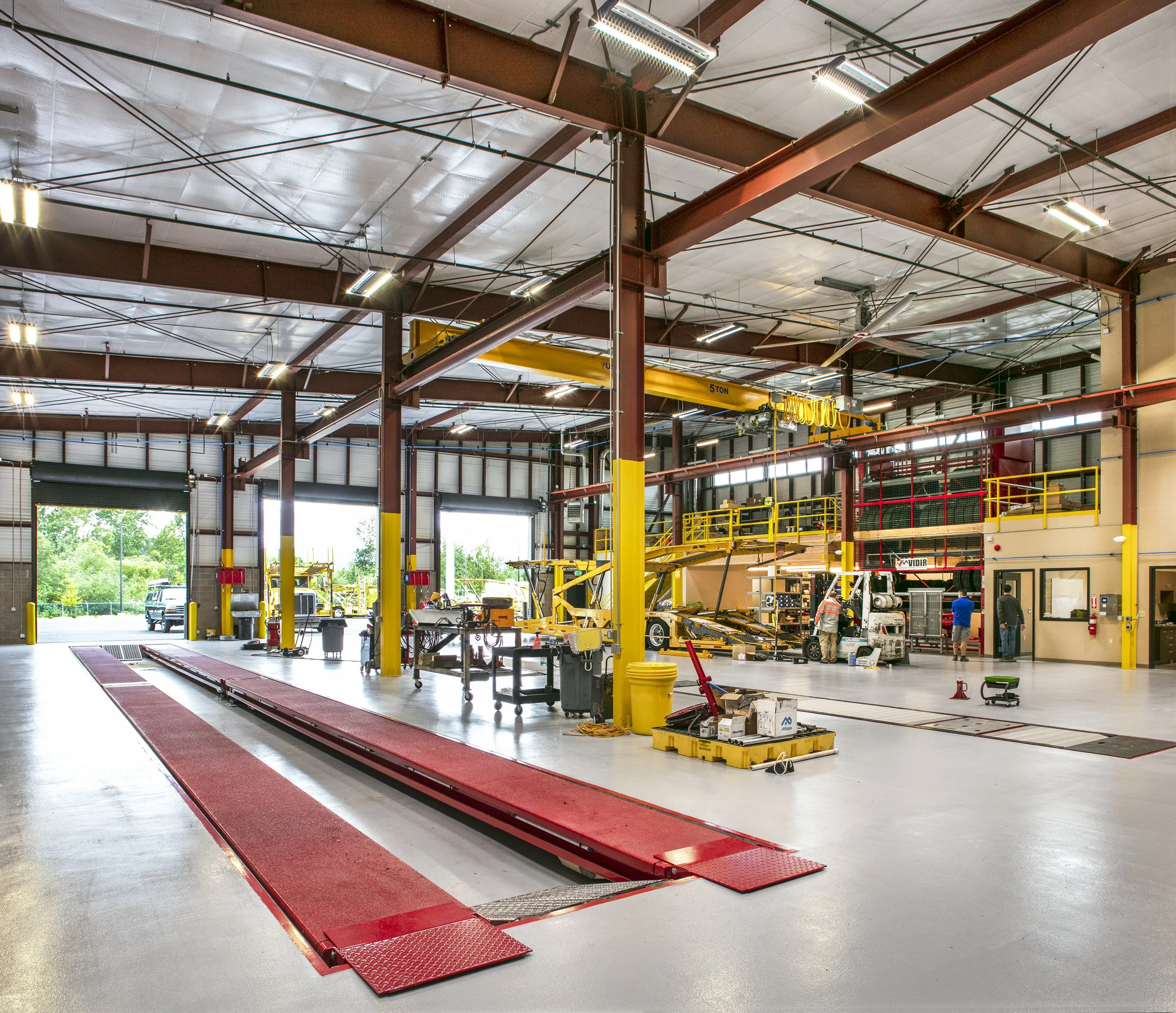Inside a large industrial workshop with a high ceiling, metal beams, and overhead lighting. There are three garage doors, some open, revealing trees and vehicles outside. The workspace includes a red vehicle lift, various tools, equipment, and a yell