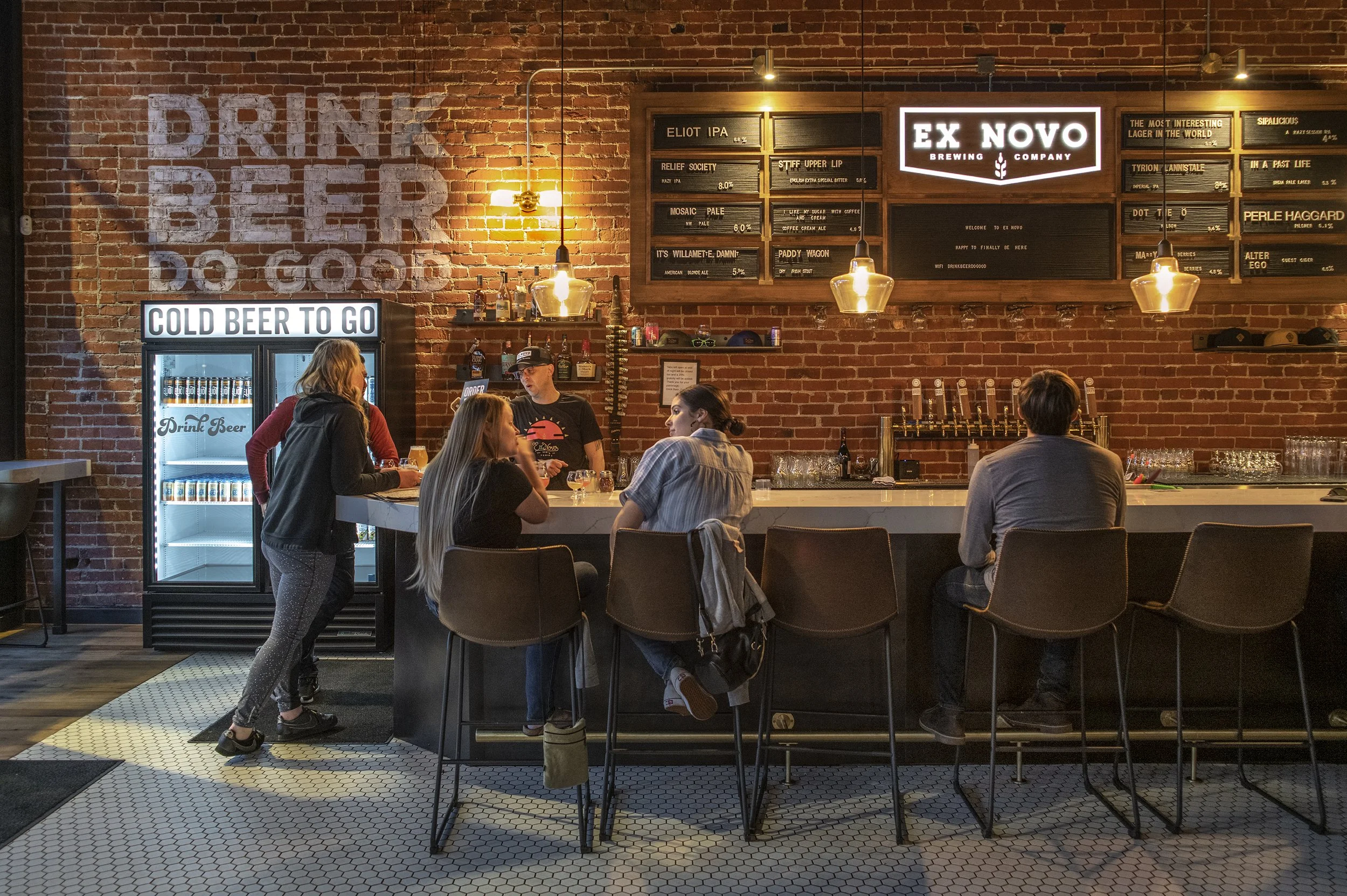 People sitting at a bar in a brewery, engaging in conversation, with a brick wall background featuring signage and a refrigerator labeled 'Cold Beer To Go'.