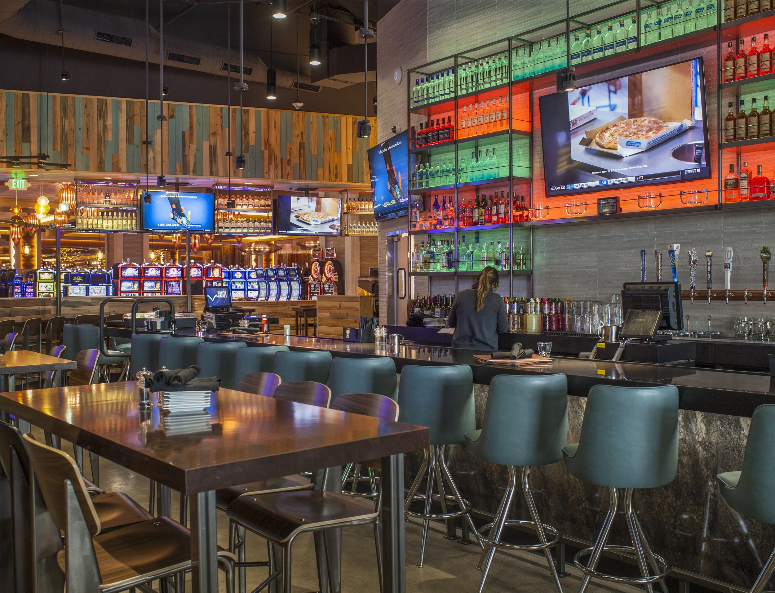 Inside a modern bar and casino, with a bar counter, teal chairs, slot machines in the background, multiple TV screens, and colorful liquor shelves.