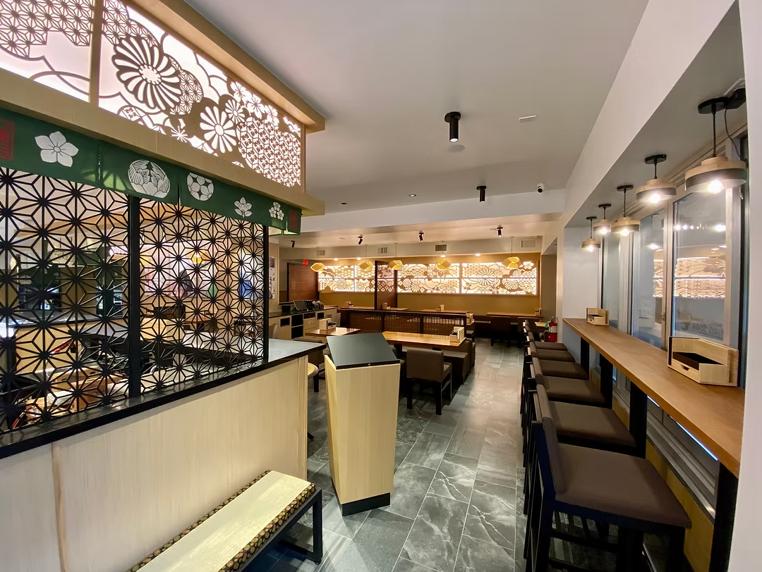 Interior of a restaurant with traditional Japanese design elements, including decorative wooden panels with floral patterns, a counter with bar stools on the right, and a seating area with tables and chairs in the background.