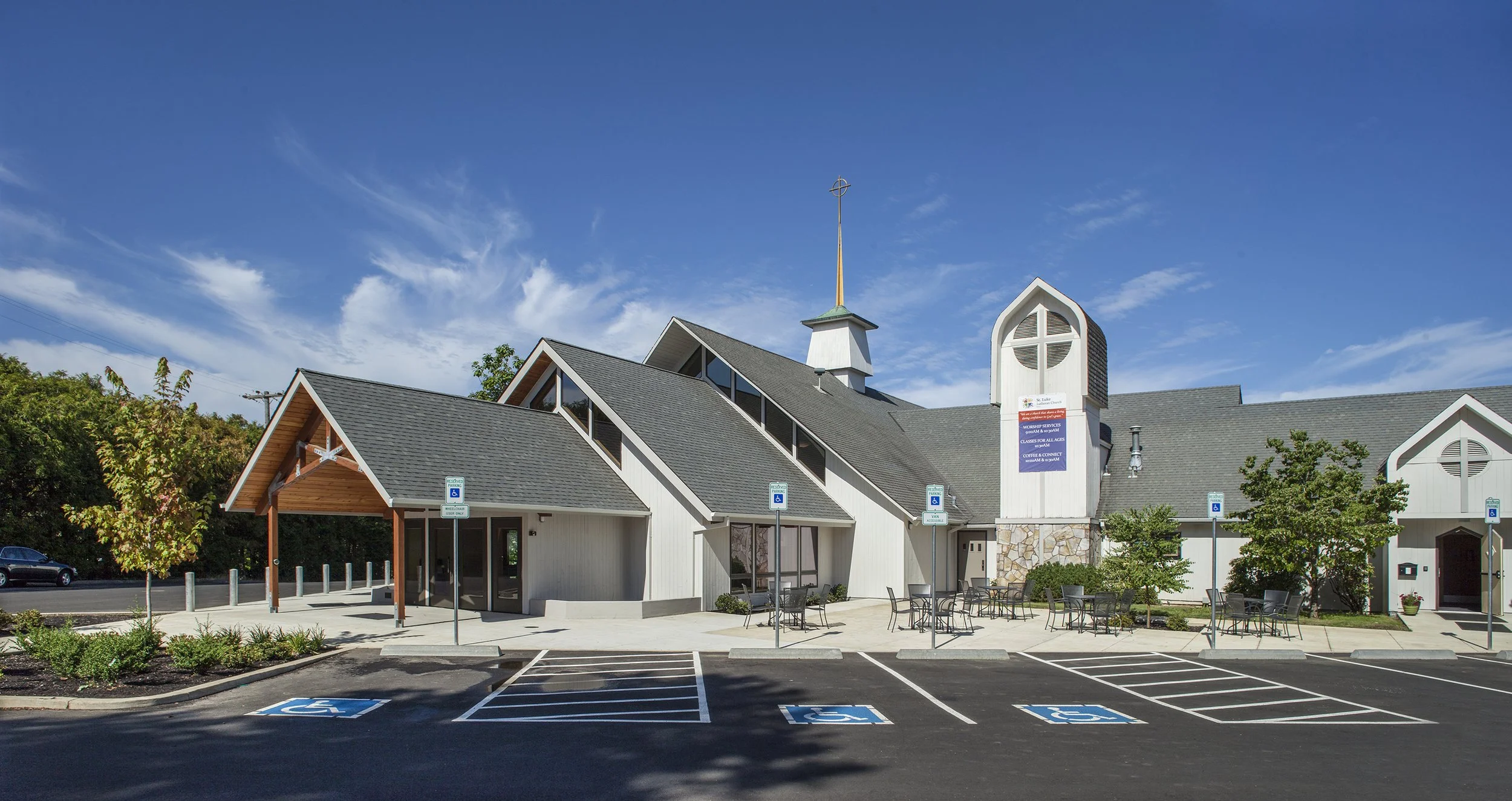 Modern church building with a parking lot and accessible parking spaces, surrounded by trees under a blue sky with some clouds.