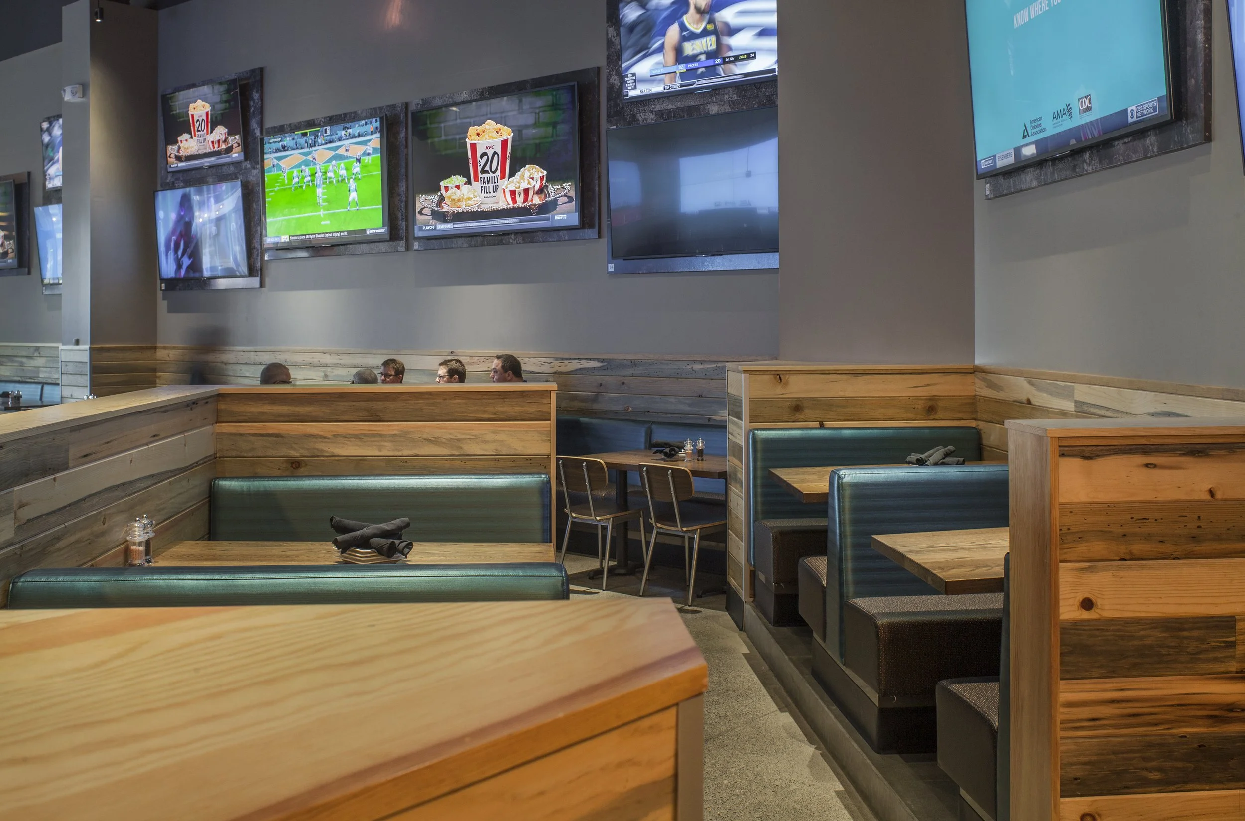 Empty booth seating with green upholstery at a sports bar or restaurant, with multiple TVs mounted on the wall showing sports and food advertisements.