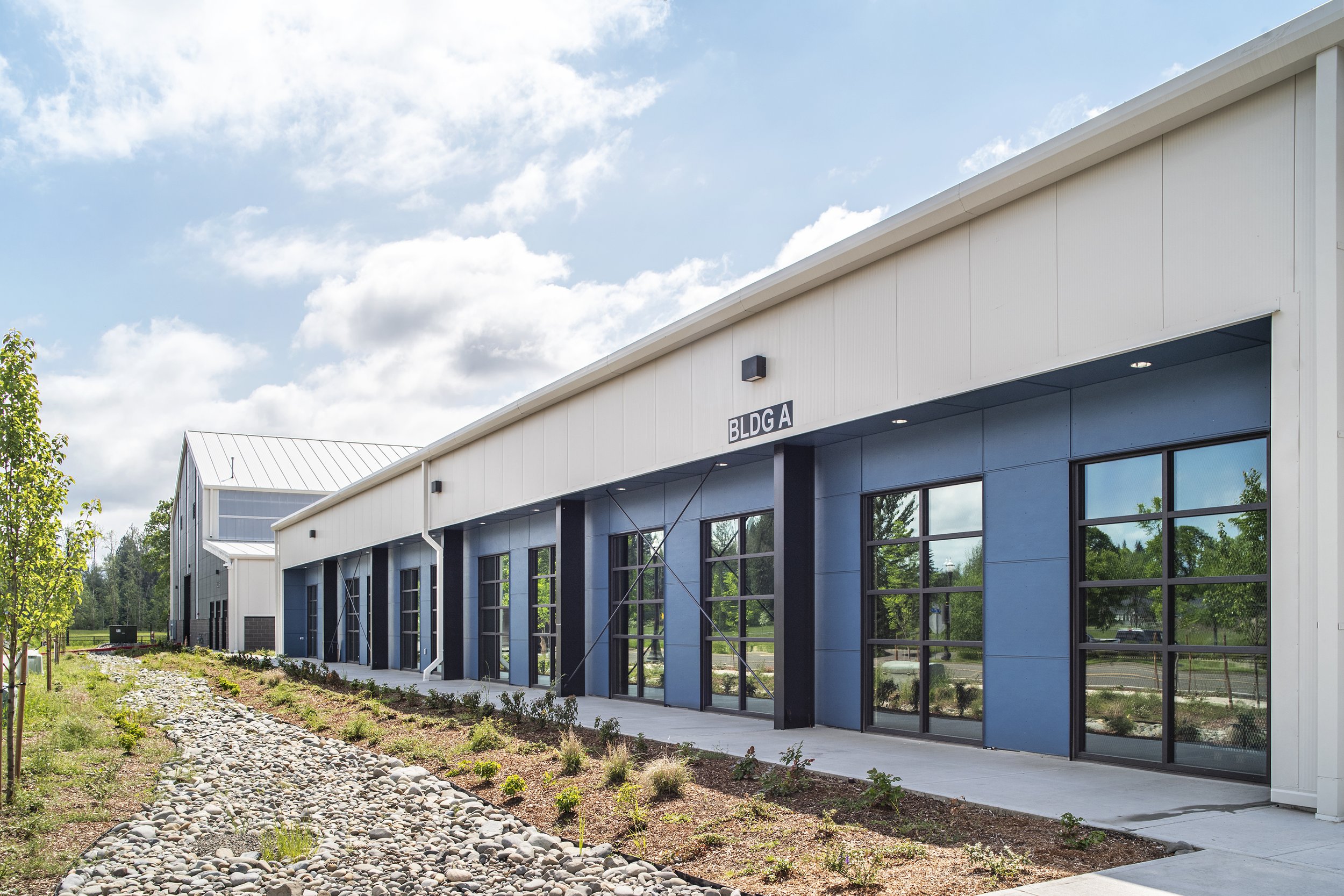 Modern commercial building with large glass doors, labeled 'BLDG A,' surrounded by landscaped area with small plants and rocks, under a partly cloudy sky.