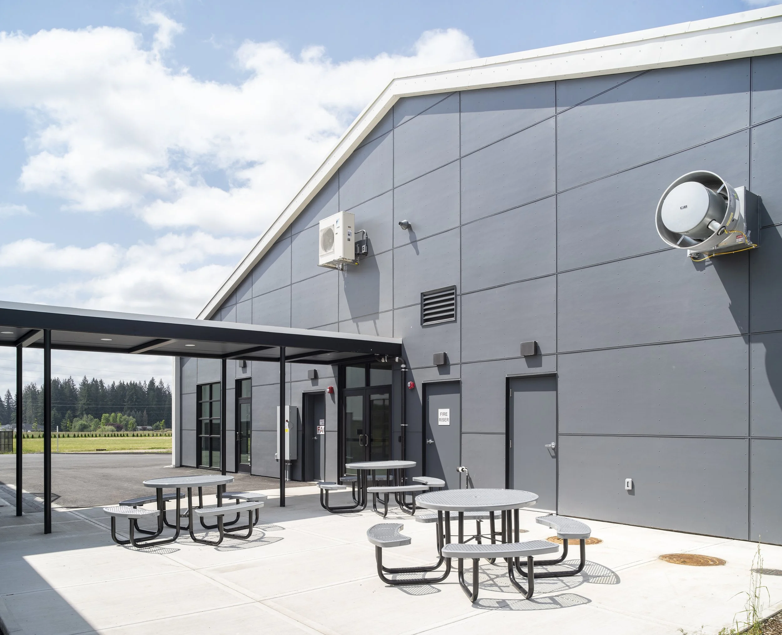 Outdoor area with gray building wall, air conditioning unit, exhaust fan, and picnic tables under a metal canopy, with a parking lot and trees in the background.