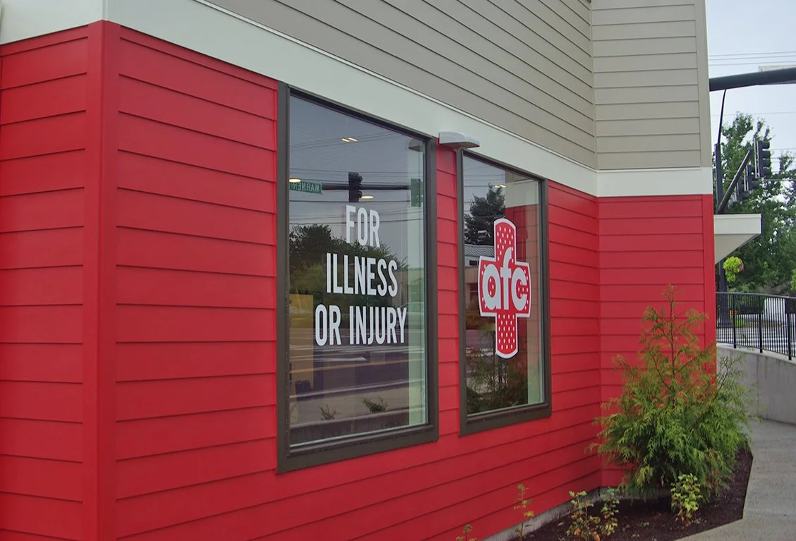 Exterior view of a red and beige building with large front windows displaying a medical cross logo with the letters "afc" and the words "For Illness or Injury".