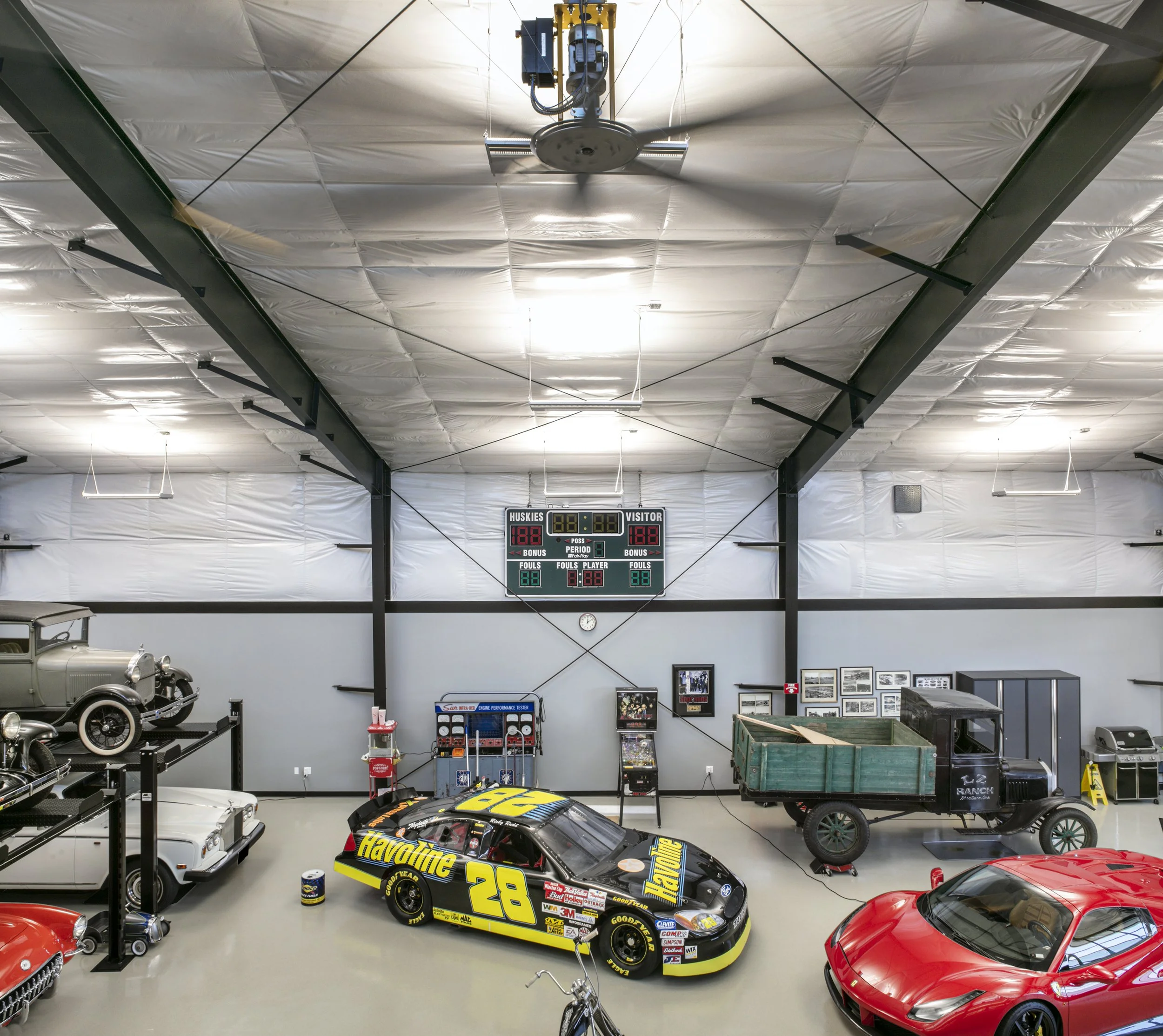 Indoor garage showcasing vintage and racing cars, including a yellow and black NASCAR stock car, a red sports car, and classic vehicles with a scoreboard overhead.