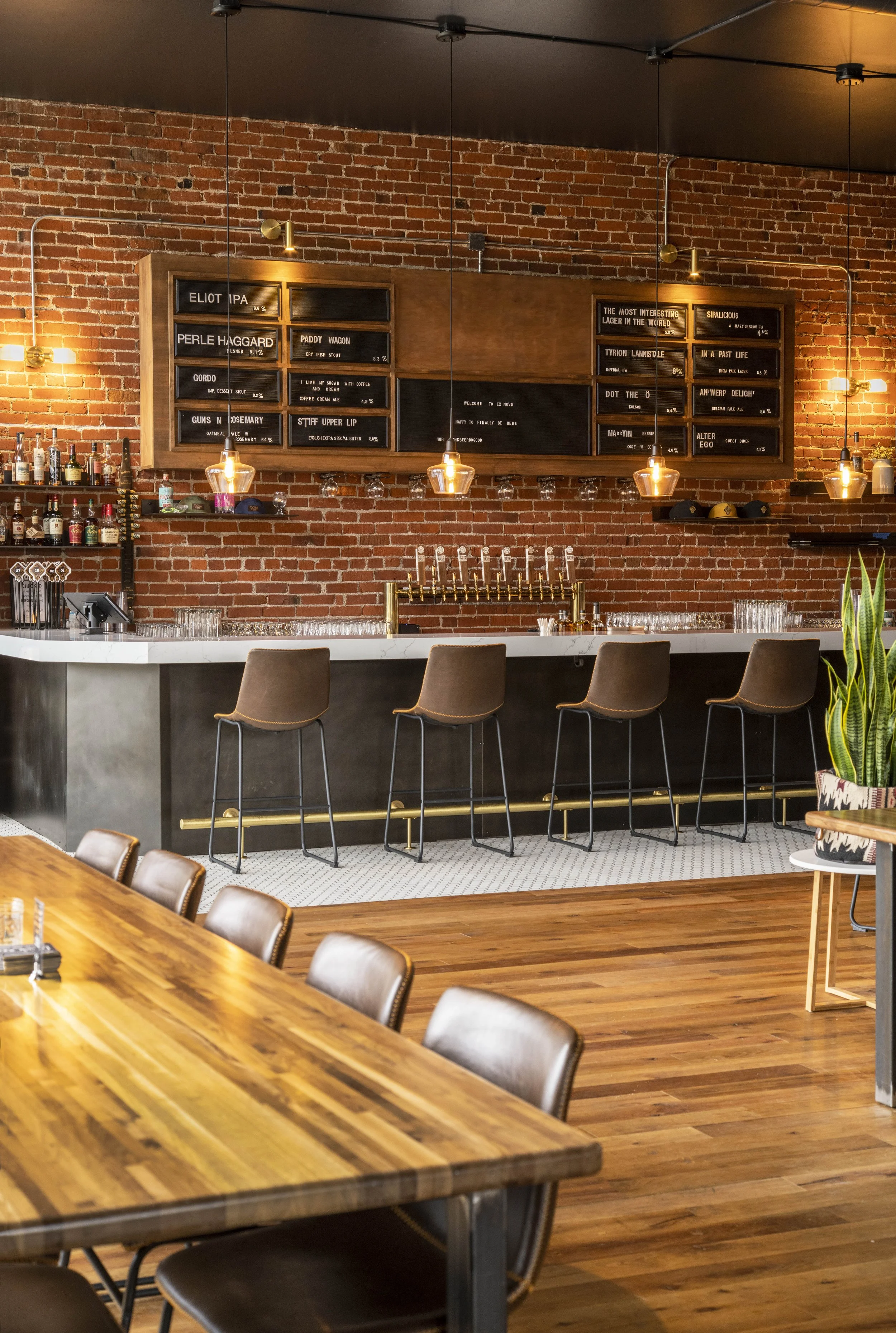 A modern bar with a brick wall, hanging pendant lights, a white bar counter with brown chairs, a wooden table with chairs, bottles of alcohol on a shelf, and blackboards displaying drink names and prices.