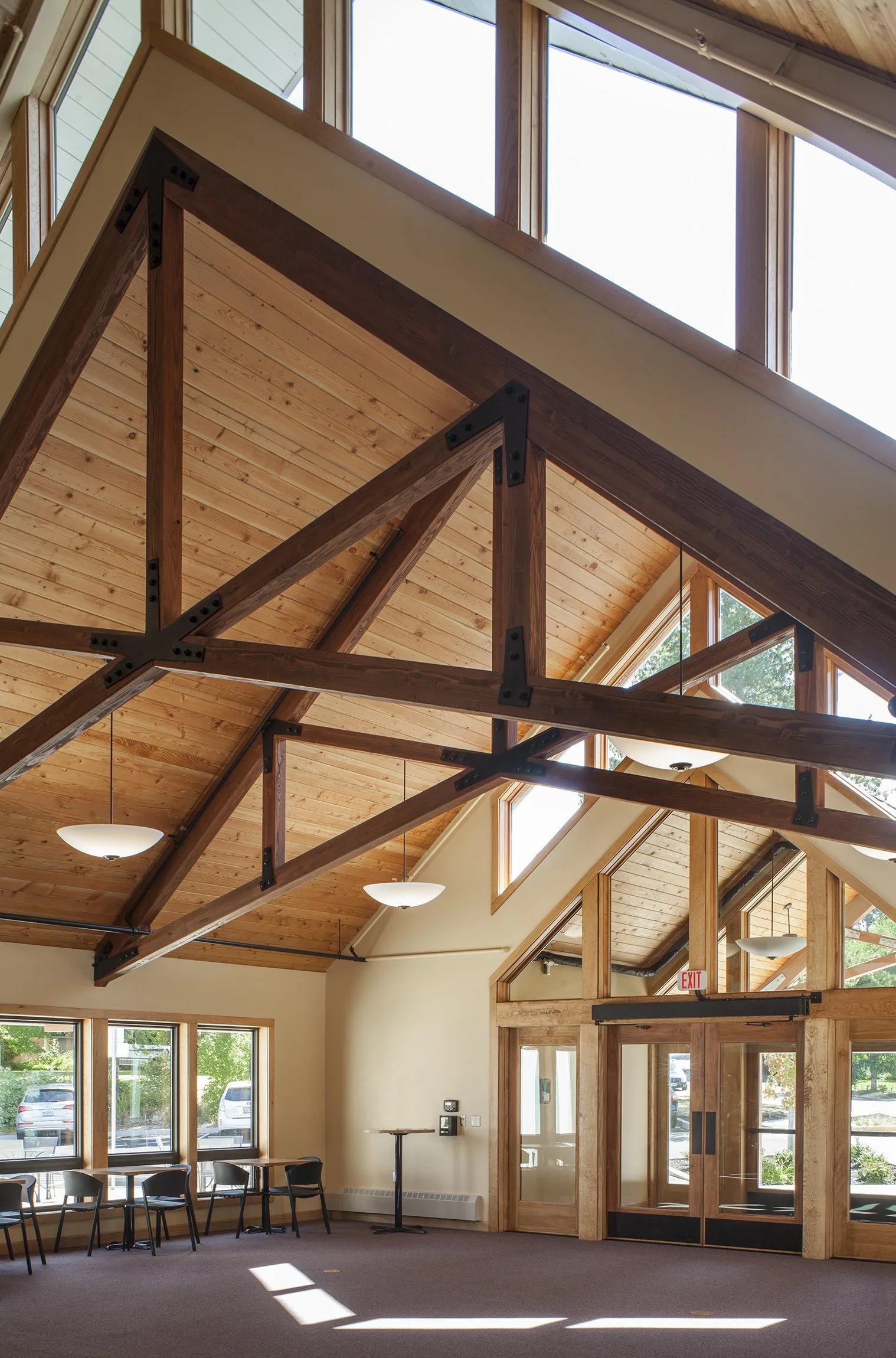 Interior of a building with a high, wood-paneled ceiling supported by exposed wooden beams, large windows, and a door with glass panels. There are chairs and tables along the windows, with sunlight casting shadows on the carpeted floor.