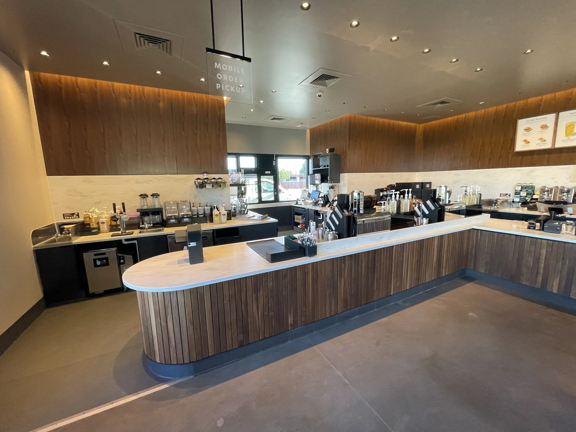 Inside of a coffee shop with a curved wooden counter, various coffee machines, and a menu board on the wall. Natural light from the front windows illuminates the space, which has a warm, modern decor.