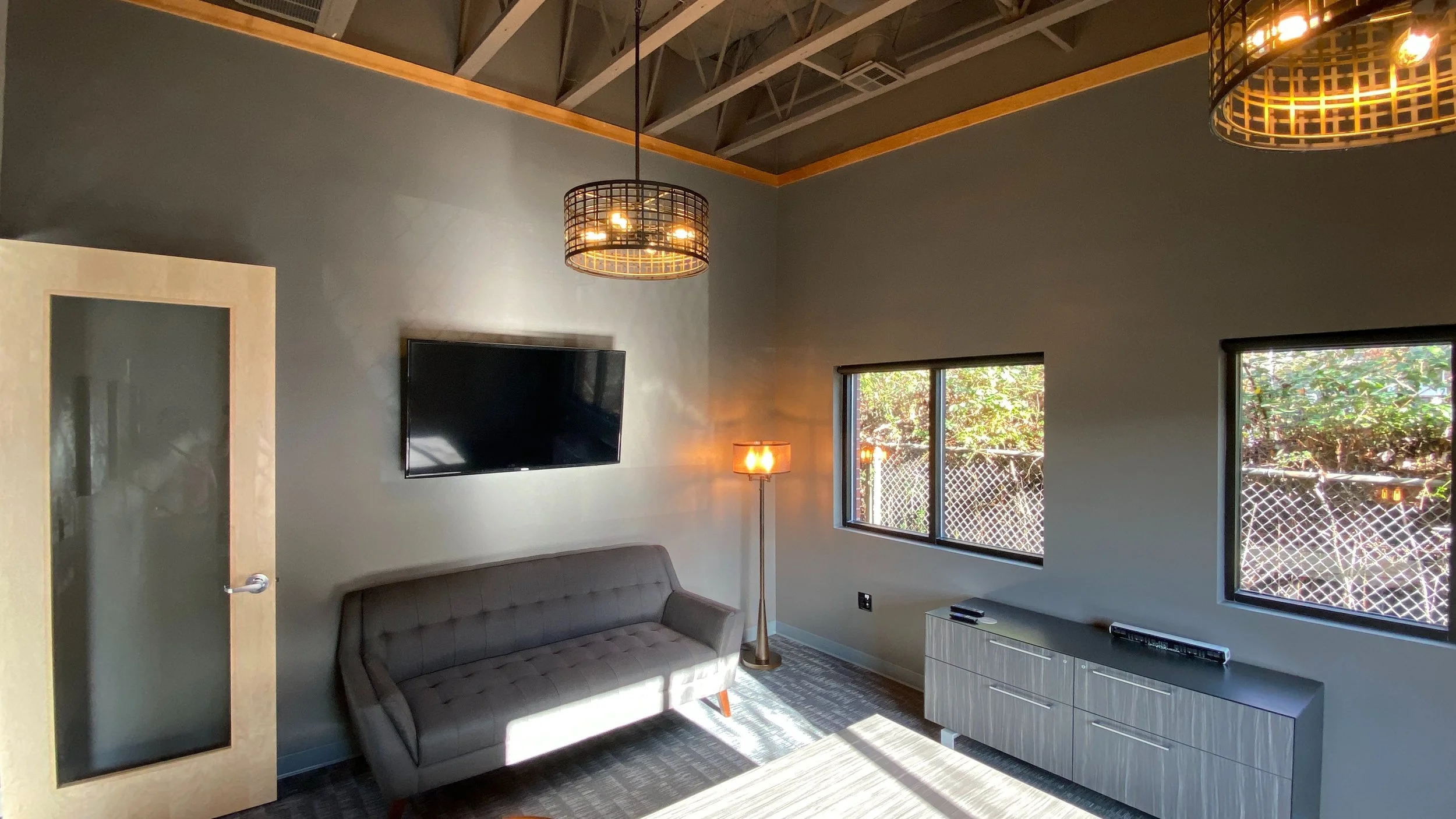 Bright living room with two windows, a mounted TV, a gray sofa, a tall floor lamp with an orange lampshade, a modern TV stand, and two ceiling pendant lights with black metal frames.