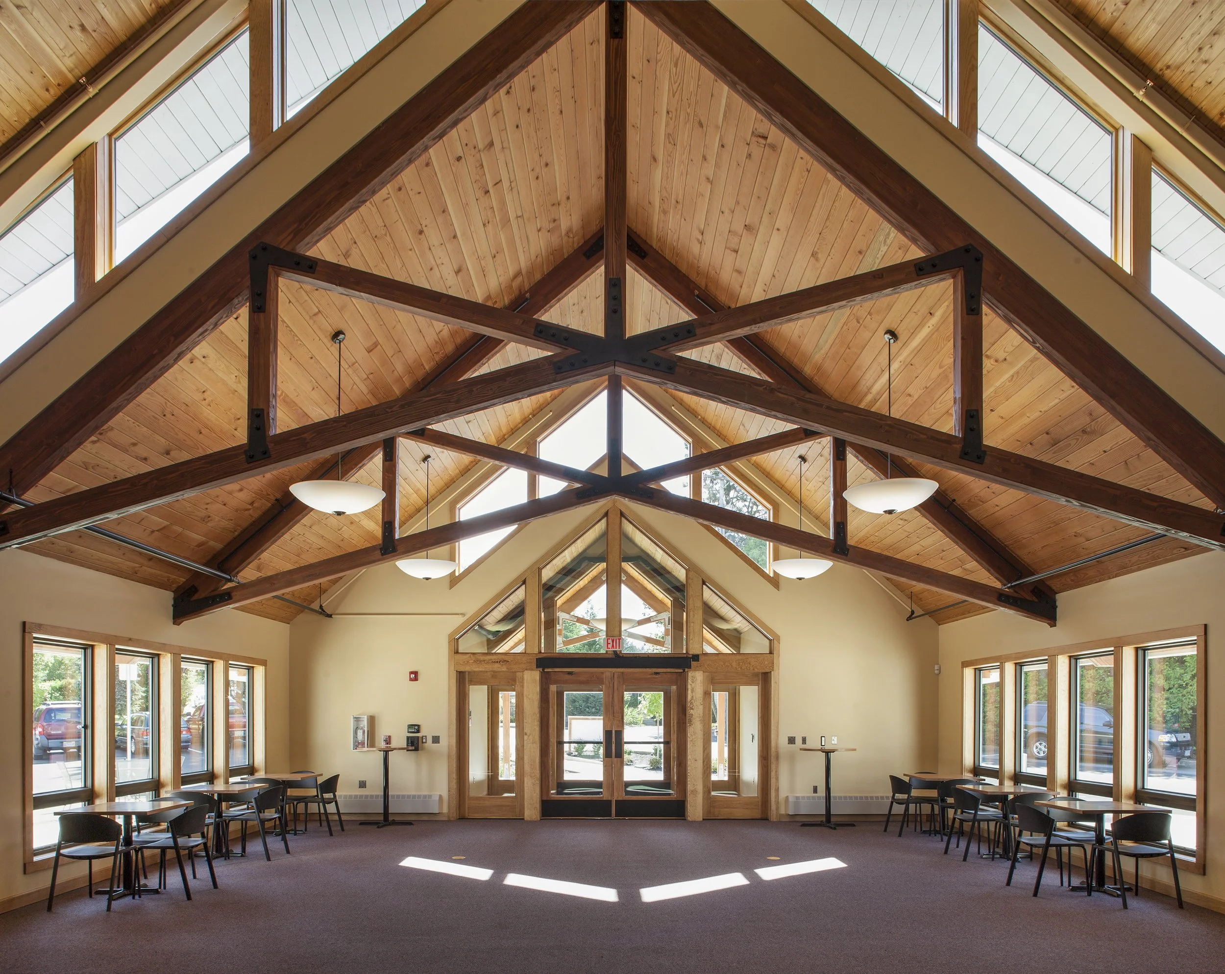 Large, open room with wooden vaulted ceiling, skylights, and large windows; wooden door with glass, chairs and tables along the walls.