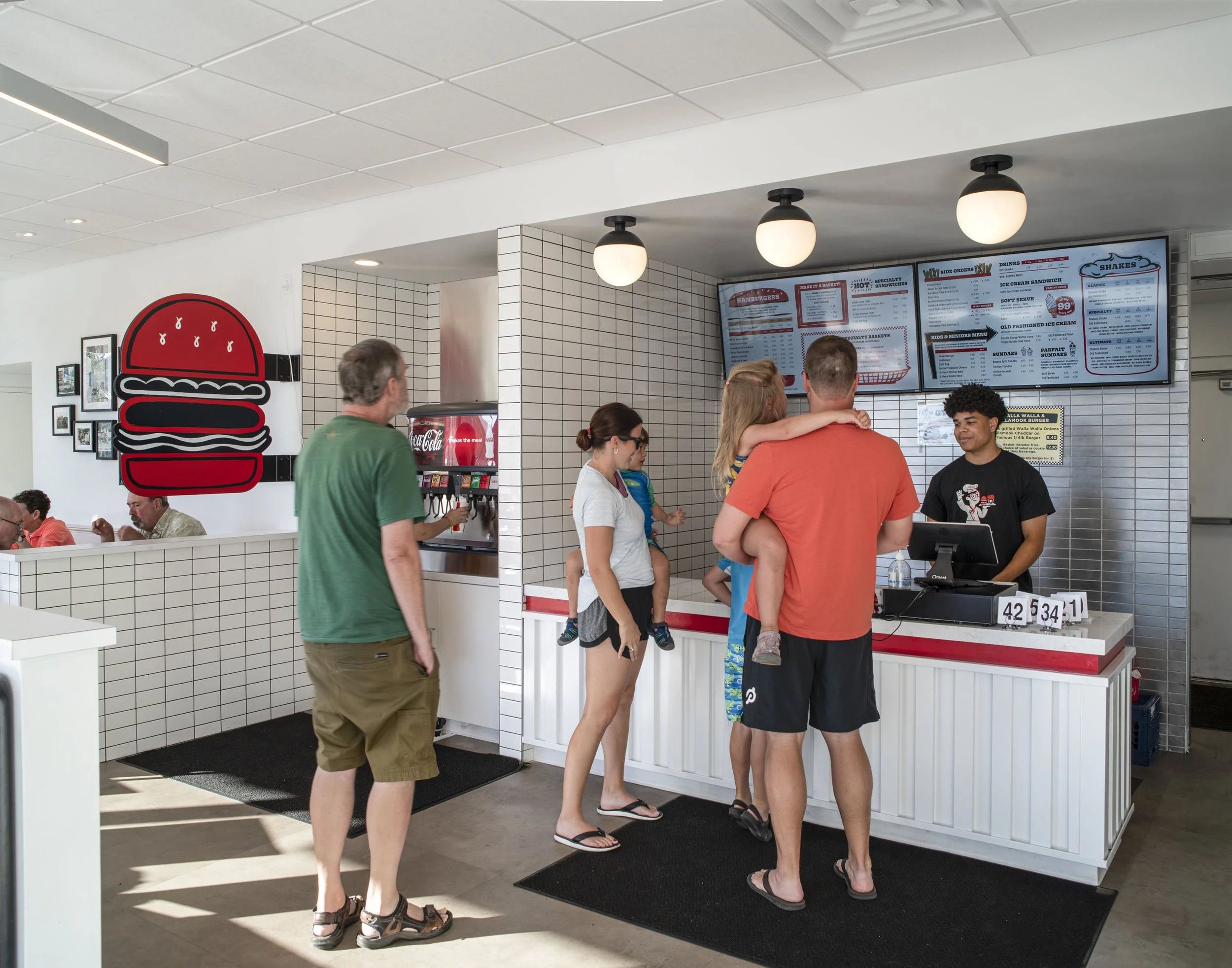 People ordering food at a fast-food restaurant counter with digital menu displays overhead, a soda fountain machine, and a large burger graphic on the wall behind.