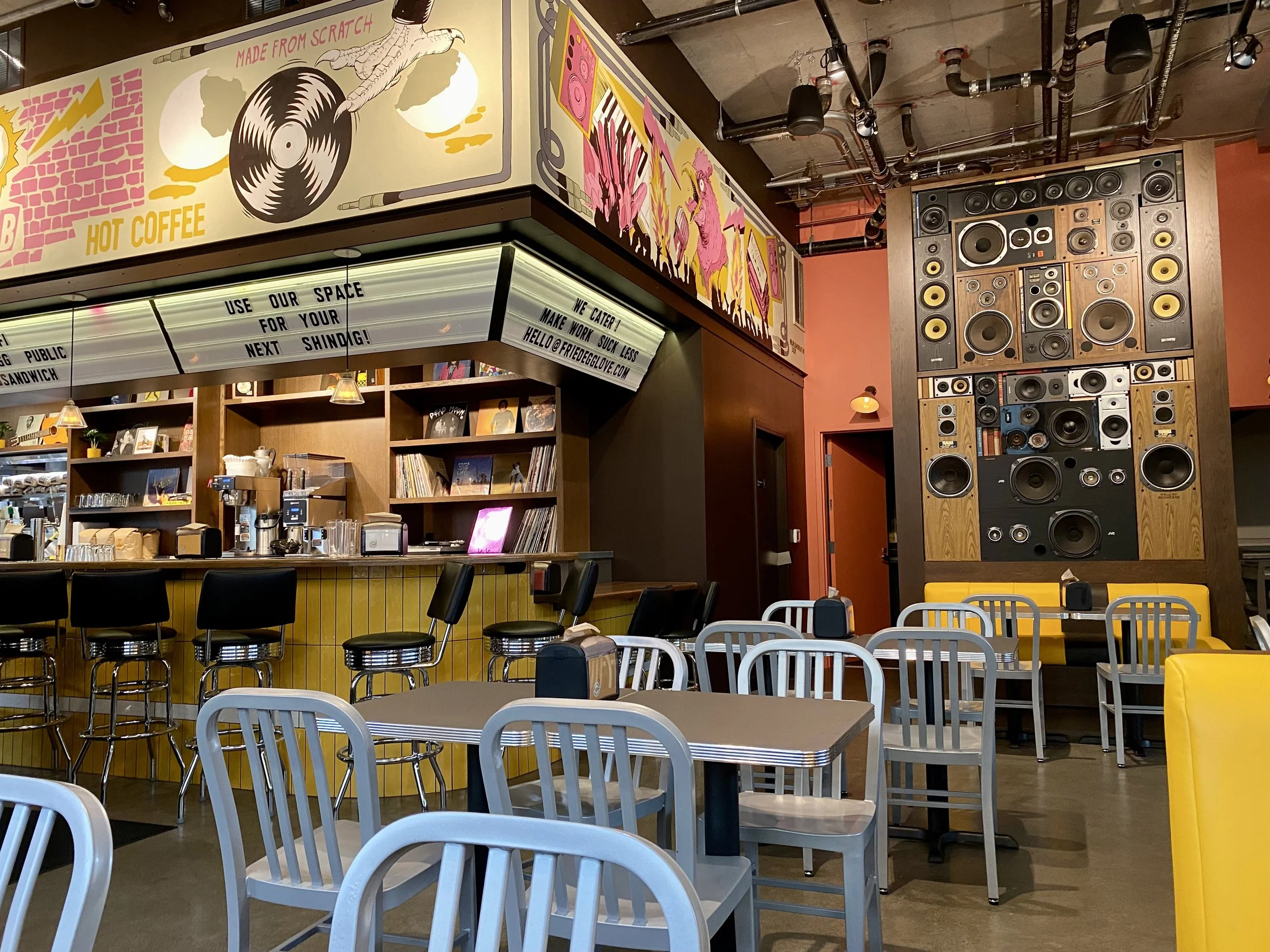 Interior of a coffee shop featuring a vibrant wall art, a bar with black chairs, a shelf with books and decor, and a large wall-mounted speaker system for music.