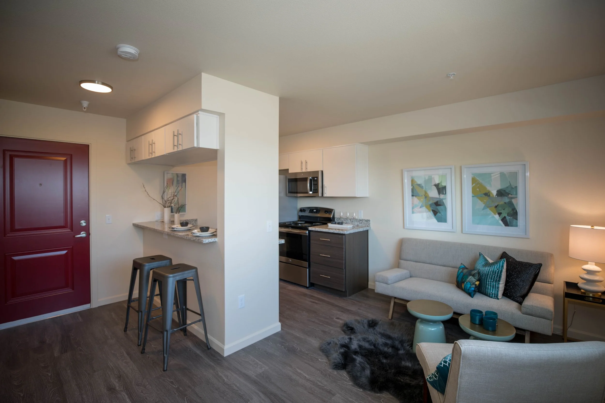 Studio apartment with a red front door, a small kitchen with white cabinets and a black stove, a cozy living area with a beige sofa, patterned cushions, a black side table with a white lamp, two abstract framed artworks, and a wood floor with a black