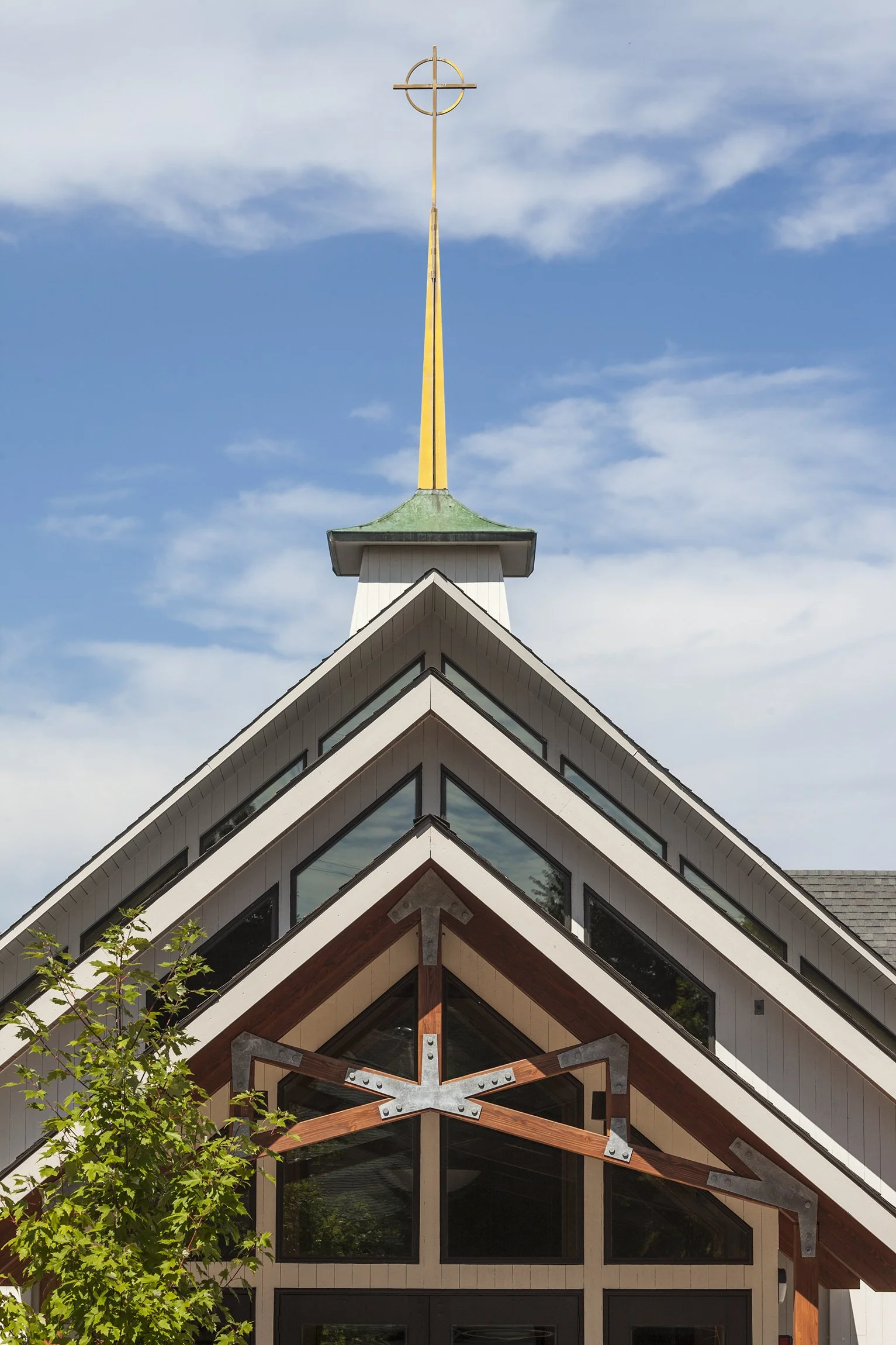 Close-up view of a church with a triangular roof, large windows, wooden and metal architectural details, and a tall cross at the top under a partly cloudy sky.