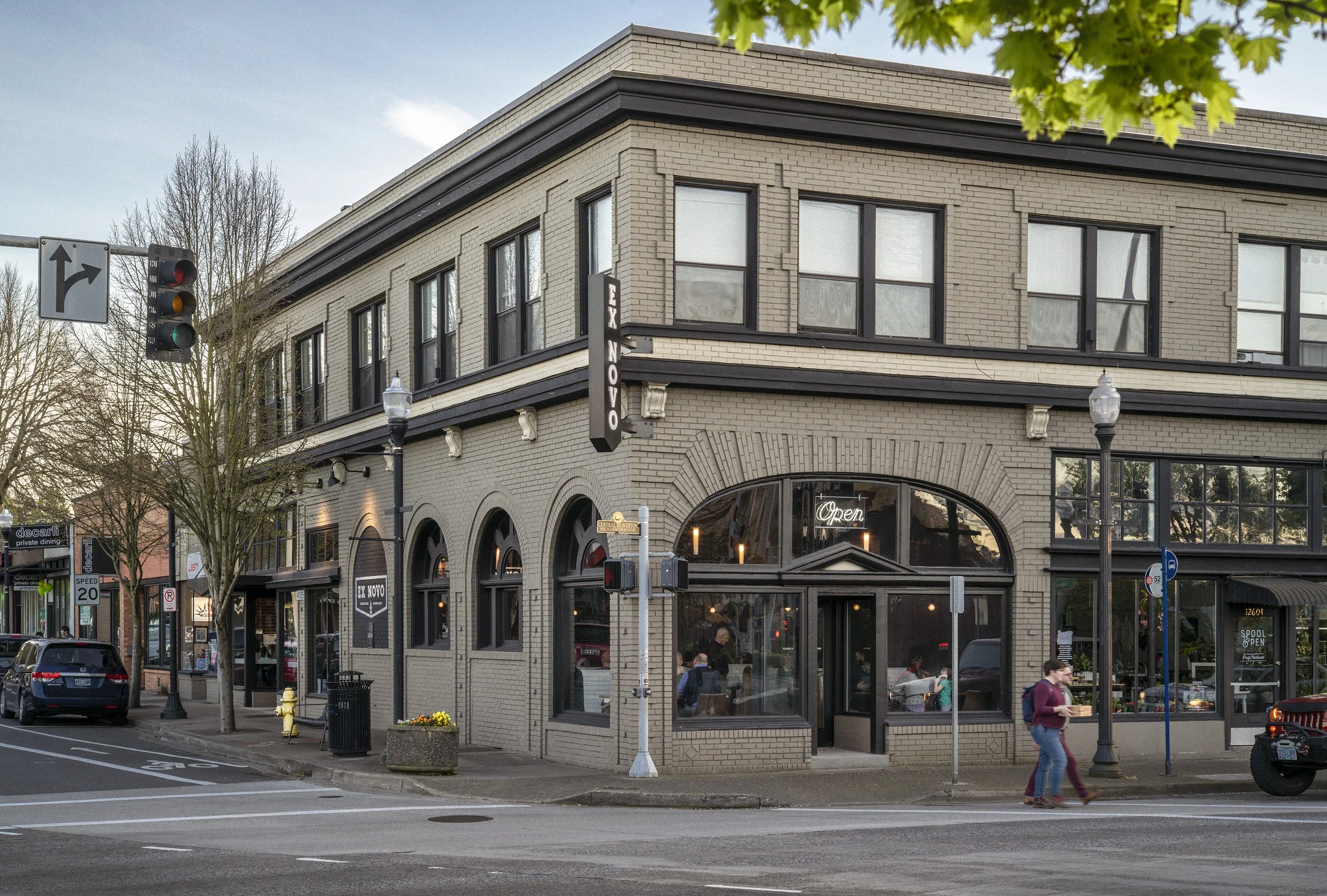 A multi-story brick building with a restaurant named 'Tex Novo' on the corner, featuring large arched windows, some with neon signs. There are streetlights, trees, a few parked cars, pedestrians crossing the street, traffic lights, and street signs i