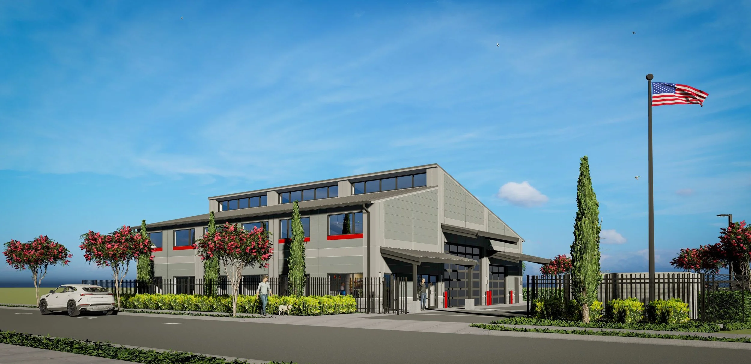 Modern industrial building with a fenced exterior, surrounded by trees with pink flowers, a parked white car on the street, and an American flag on a tall flagpole against a blue sky with a few clouds.