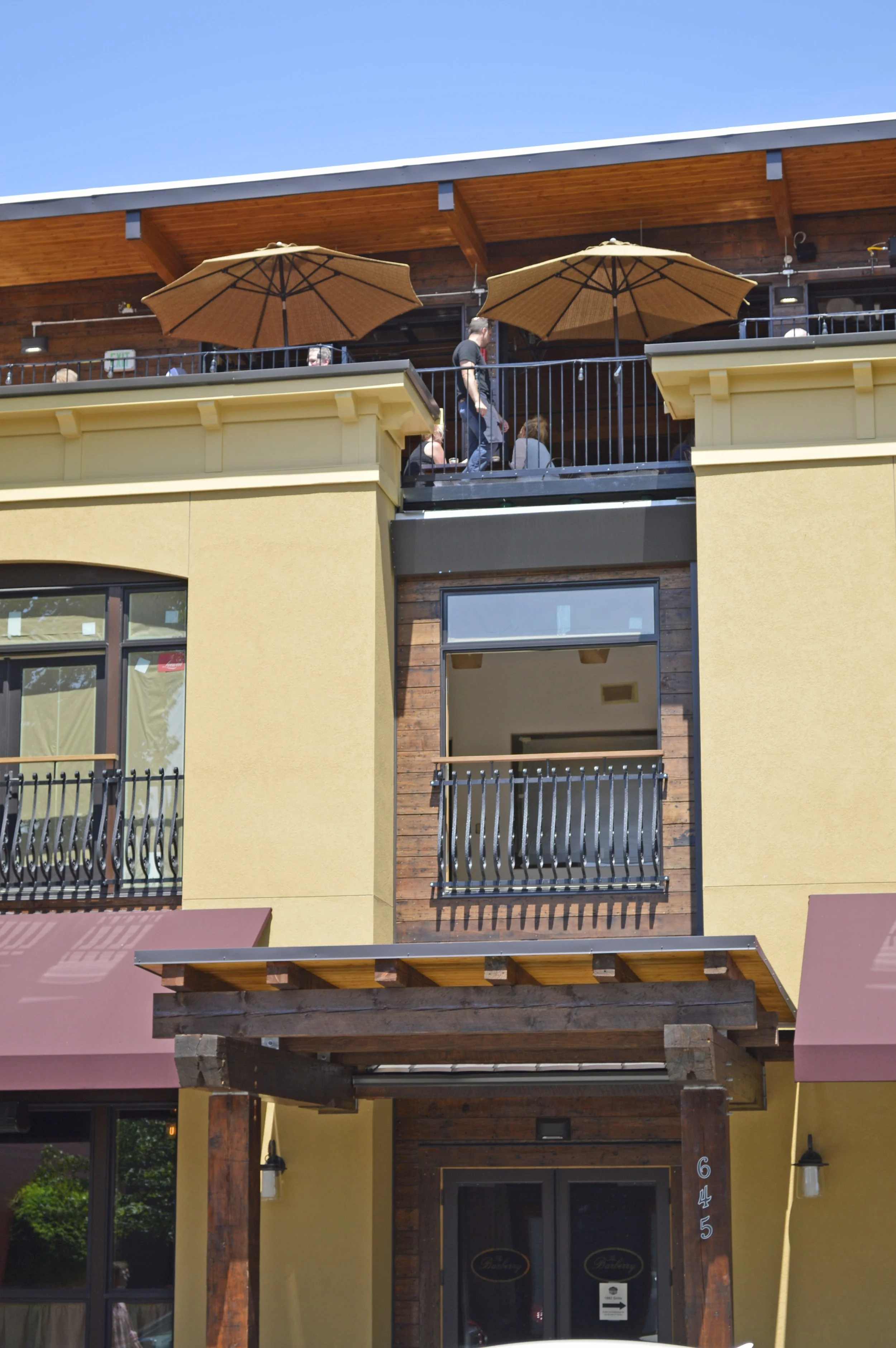 A multi-story building with outdoor patios, umbrellas, and several people sitting and walking on the upper balcony, with clear blue sky in the background.