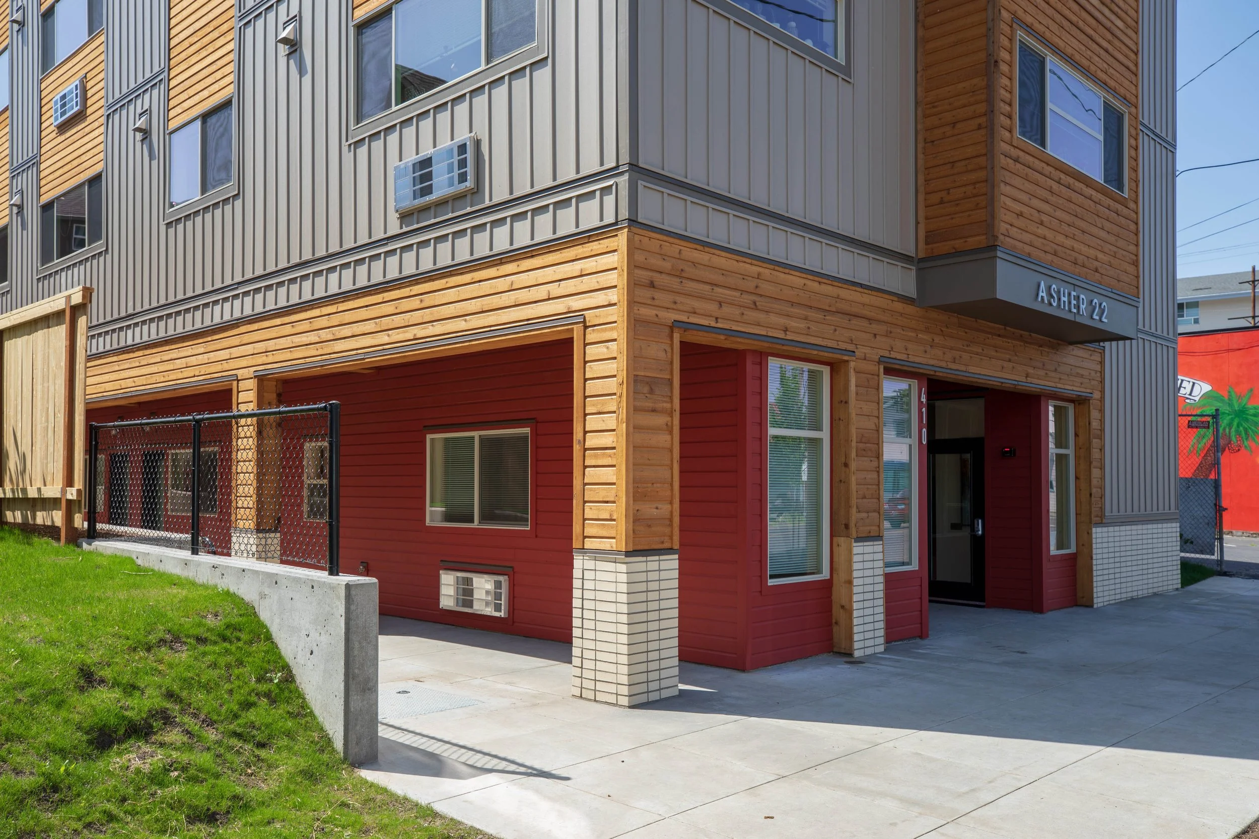 A modern multi-story building with a mix of wood and metal siding, featuring multiple windows, a small air conditioning unit, and the address 'Asher 22' above the entrance. There is a sidewalk and a small grassy area enclosed by a black chain-link fe