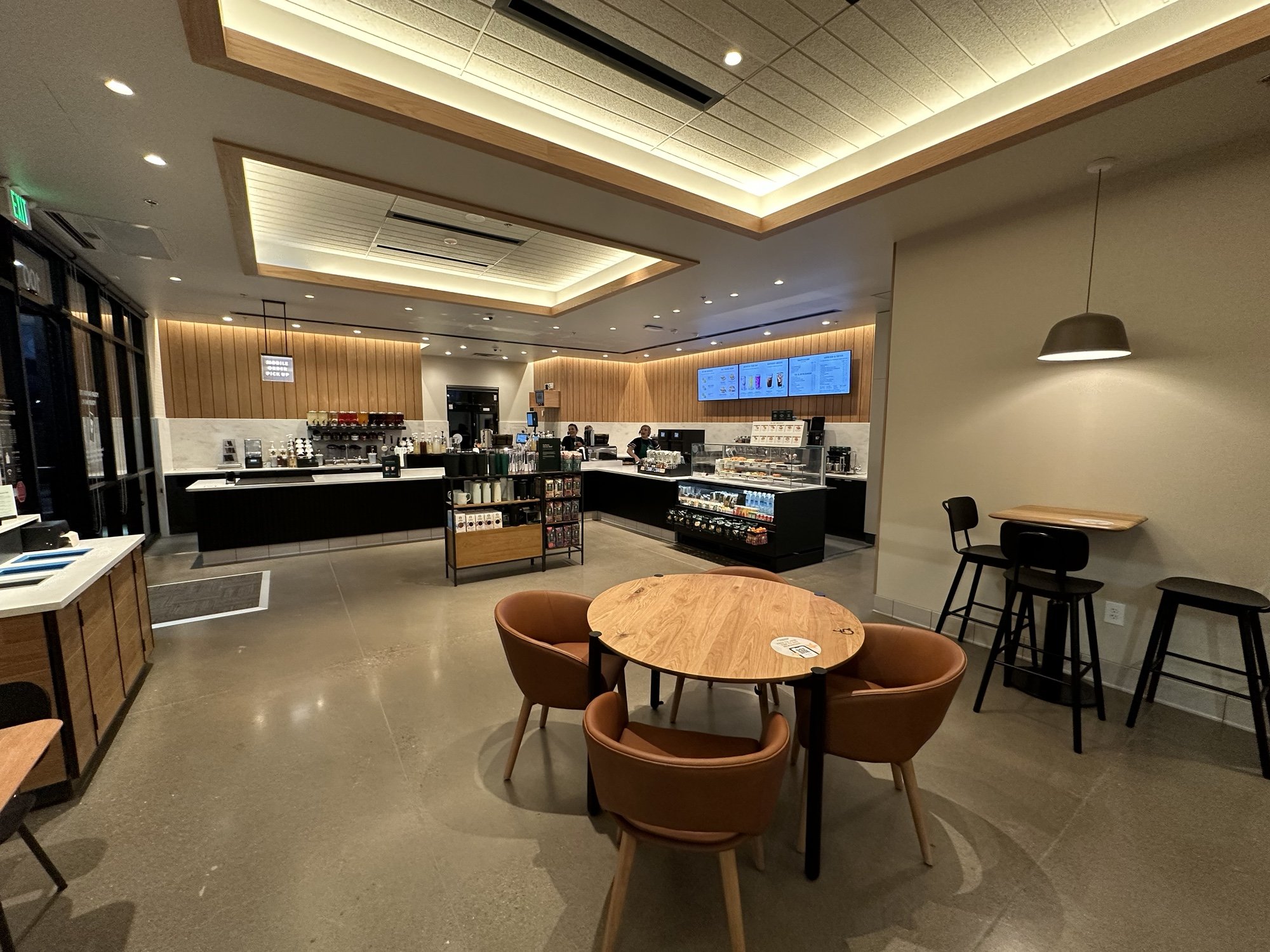Interior of a modern Starbucks cafe with a seating area, round wooden table with four chairs, a small high table with barstools, and a counter with coffee machines and menu screens in the background.