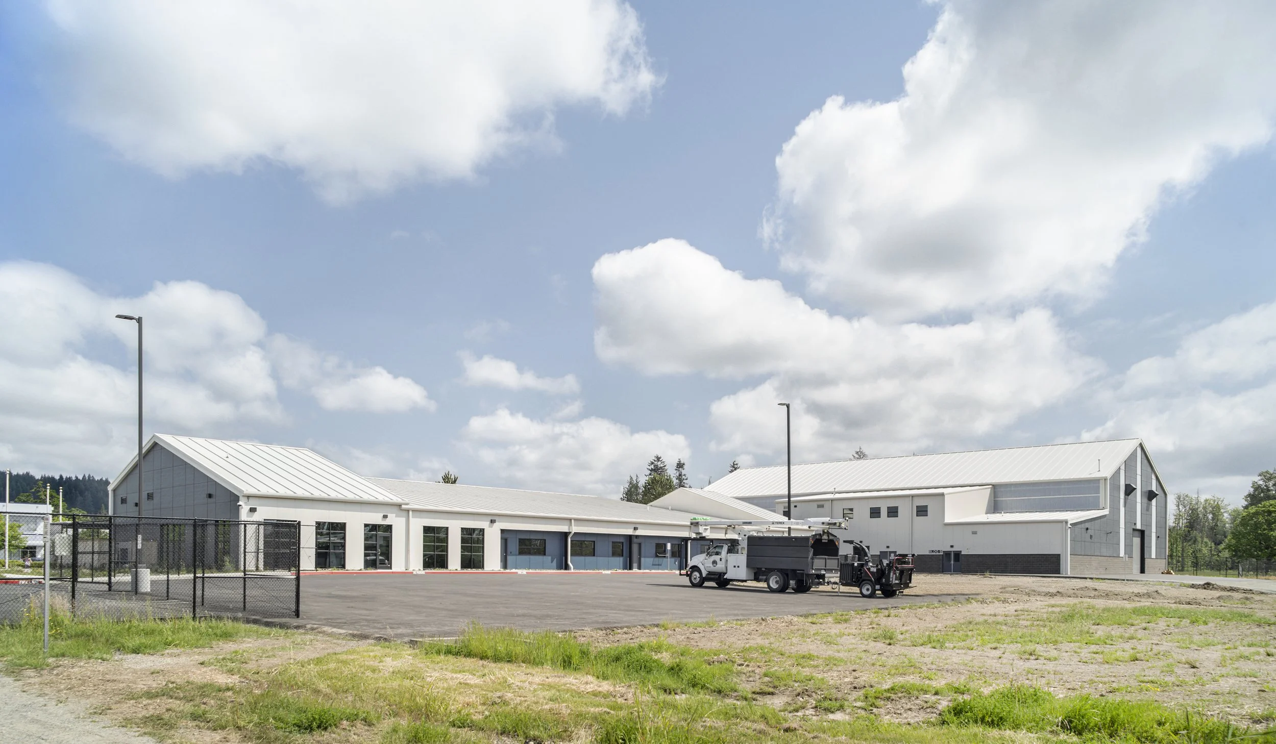 Large metal building with white walls and metal roof, surrounded by an empty parking lot and a fenced area, with utility vehicles parked nearby. Some grassy patches in the foreground, trees and a blue sky with clouds in the background.