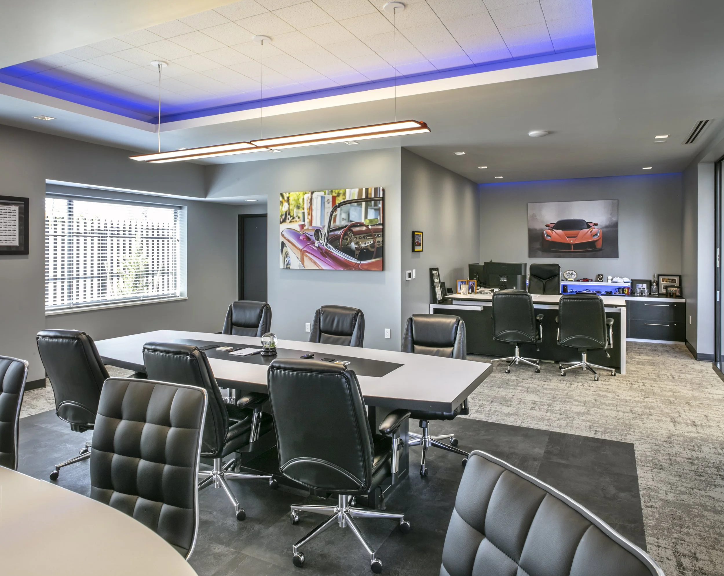 Modern office conference room with a large white table, black leather chairs, artwork of luxury cars on the walls, and a desk with computers and office supplies.