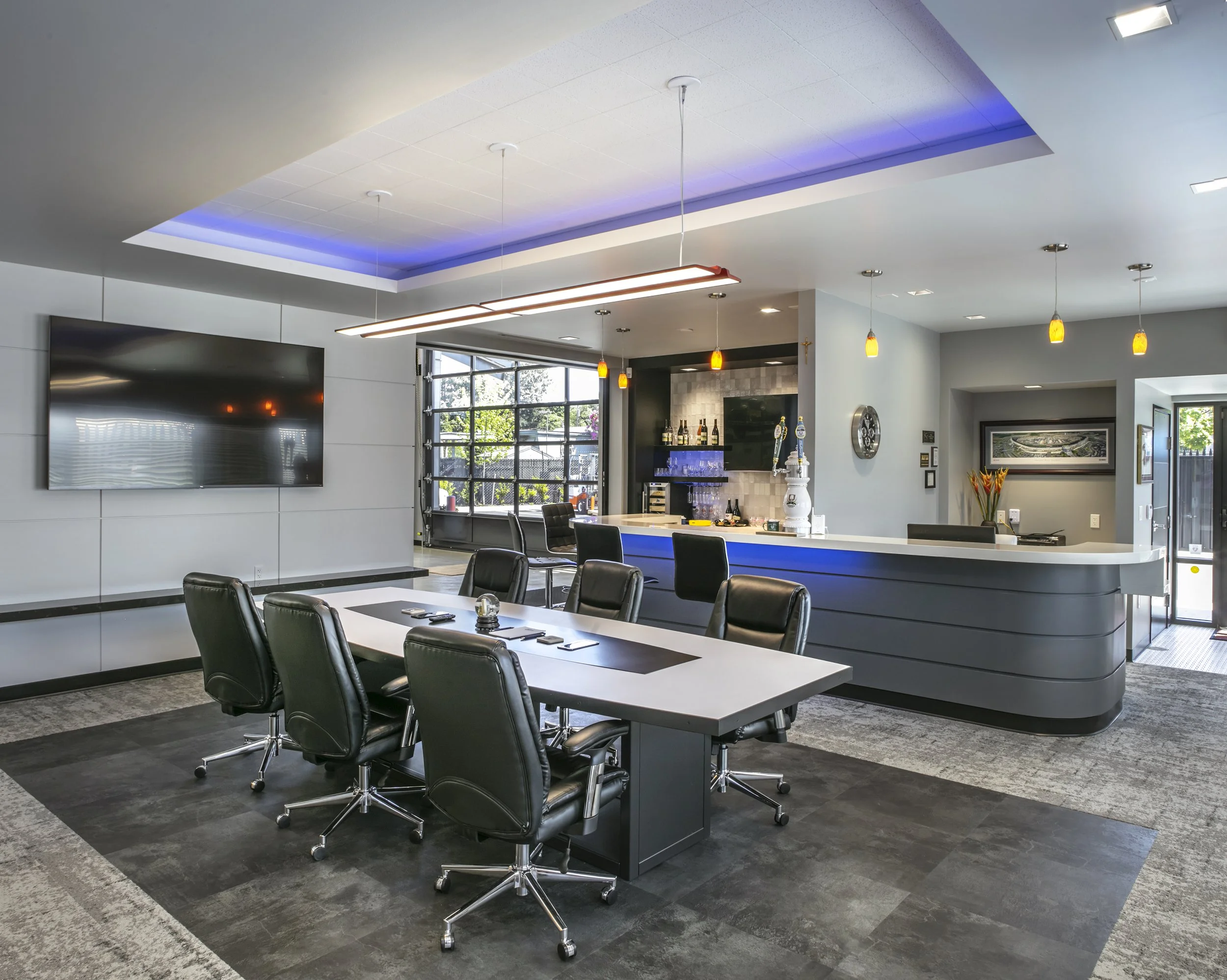 Modern conference room with a long table surrounded by black leather chairs, a large flat-screen TV on the wall, a reception bar with hanging pendant lights, and a large window letting in natural light.