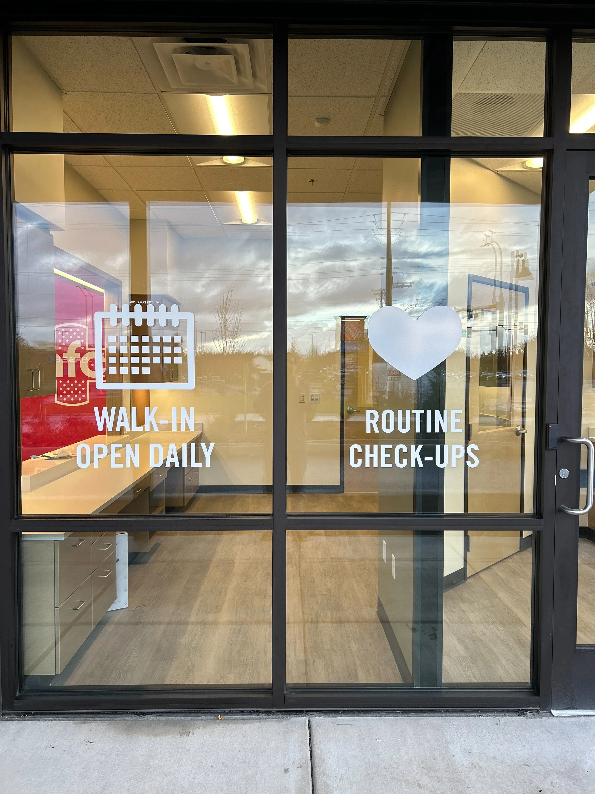 Glass storefront door with signage indicating walk-in service open daily and routine check-ups, featuring a graphic of a calendar and a heart.