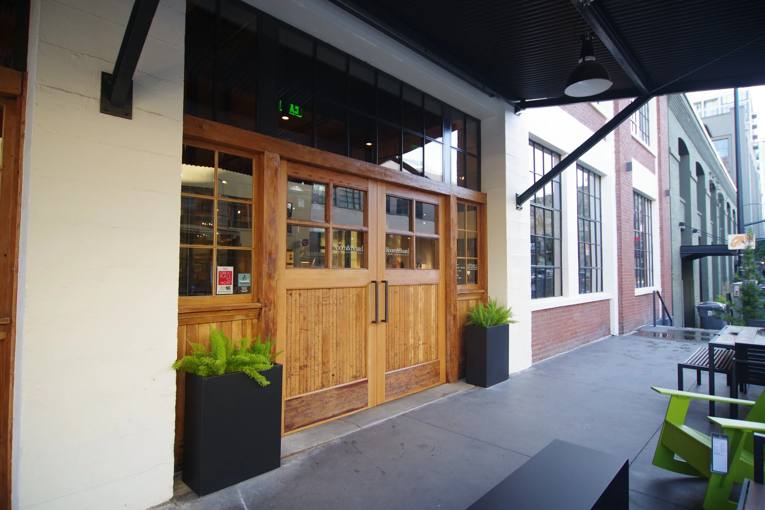 Exterior view of a building with wooden double doors, black planters with green ferns on either side, large windows, and outdoor seating including black chairs and a lime green chair, in an urban setting.